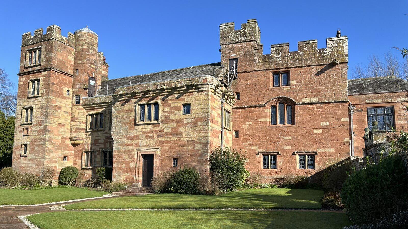 A general view of the entrance to Dalston Hall. It's a grand stone multi-storey building with a lawn and a pathway outside.