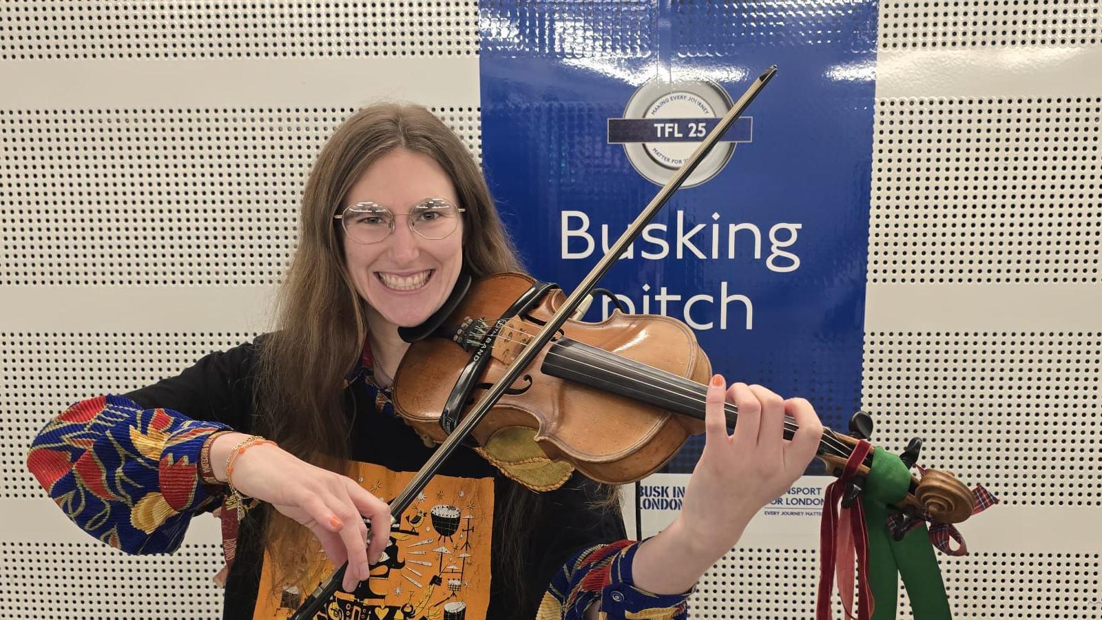 Natalie Russo, a woman with long hair and a colourful top holding a violin decorated with red and green bows