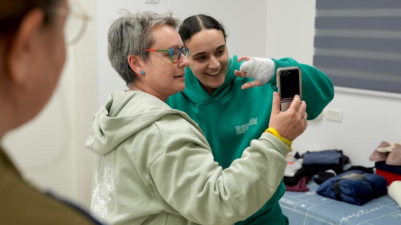 Mandy, wearing a hoody, holds up her phone during a video call as she stands with her daughter Emily, who is also wearing a hoody and a bandage around her hand