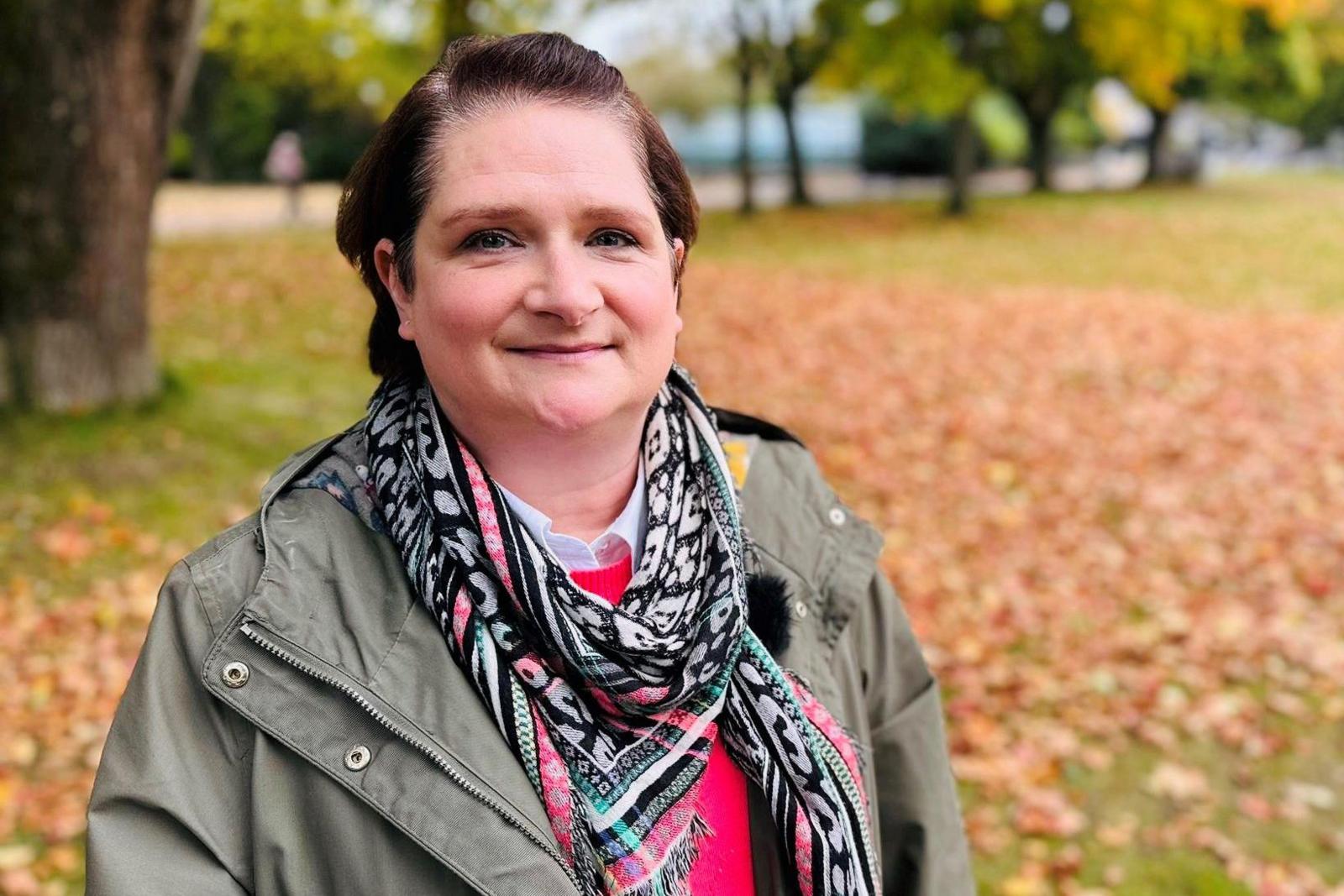 Christy Esslemont, woman wearing green jacket and scarf, standing among leaves on ground in a park, with trees in background