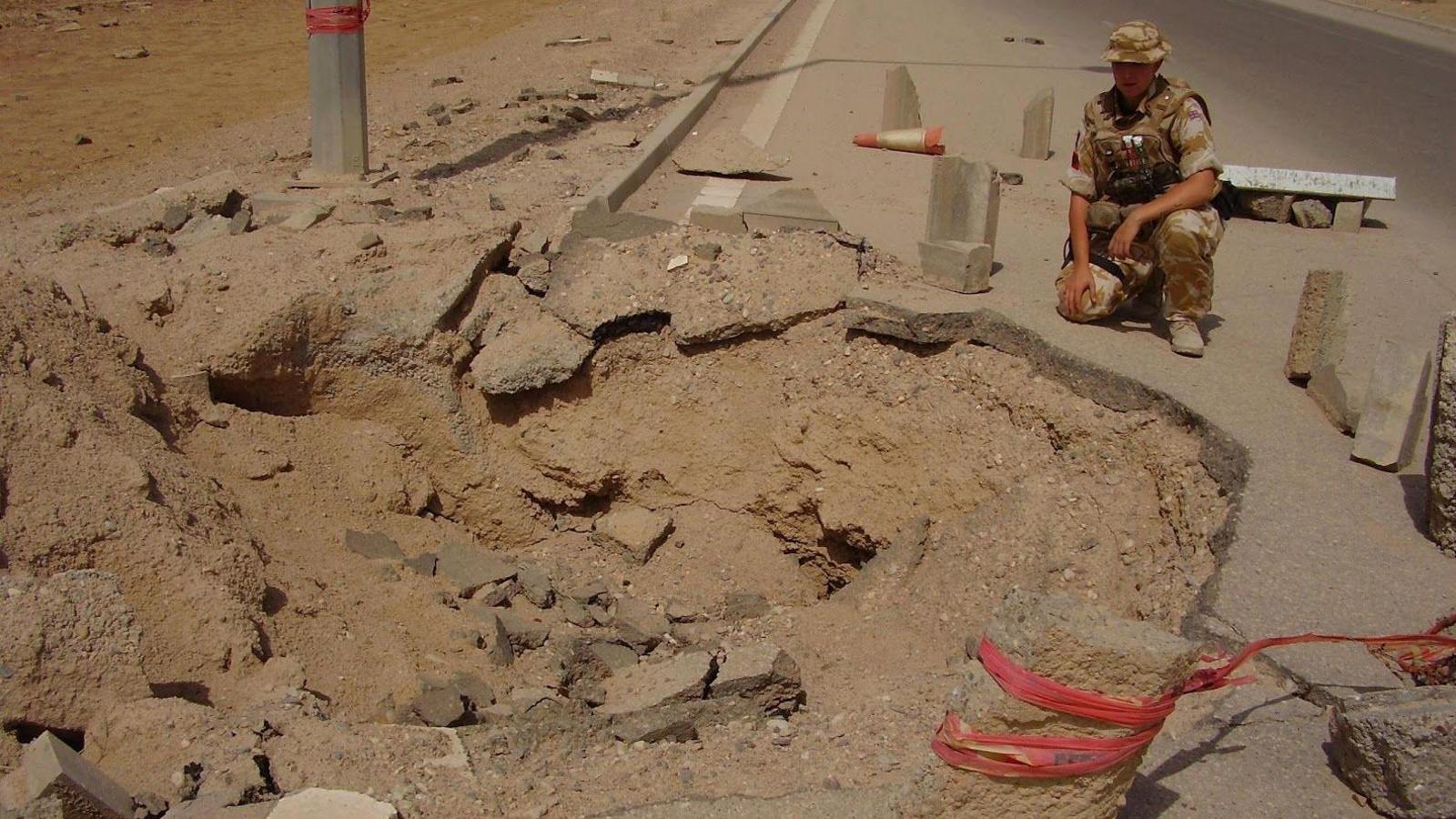 Stacey is knelt down at the side of a crater in a road looking into the crater, she is wearing desert camouflage and body armour.
