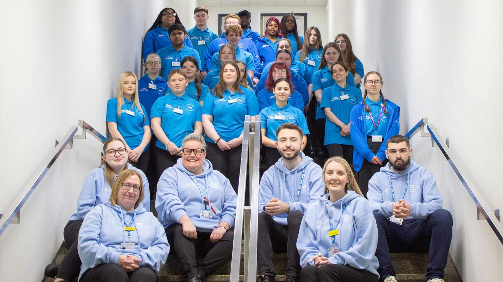 A group of staff and volunteers sit in a stairwell - the staff in light blue hoodies, and the youth volunteers in royal blue polo shirts and hoodies.