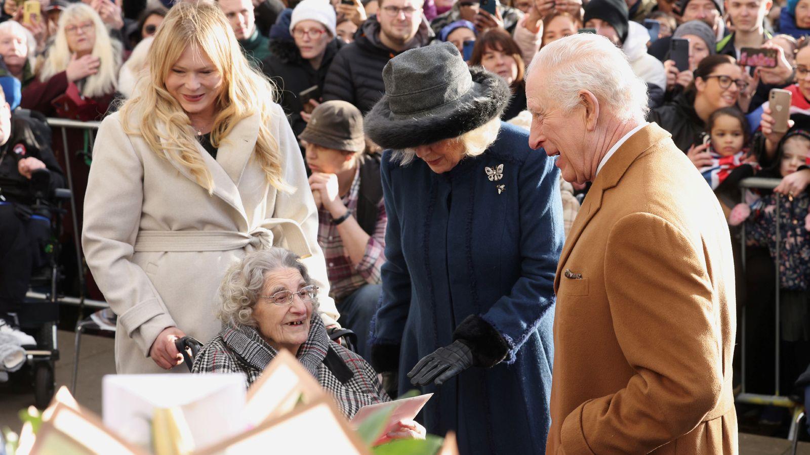 Samantha McCormick, a woman with long blonde hair and wearing a cream, woollen coat, pushes a wheelchair in which is 100-year-old Rona Grafton. She has permed grey hair and wears glasses and a check coat. Queen Camilla - wearing a black fur-rimmed hat and blue overcoat - and King Charles - in a deep tan coloured coat - are talking and laughing with Rona. In the background is a large crowd of onlookers.