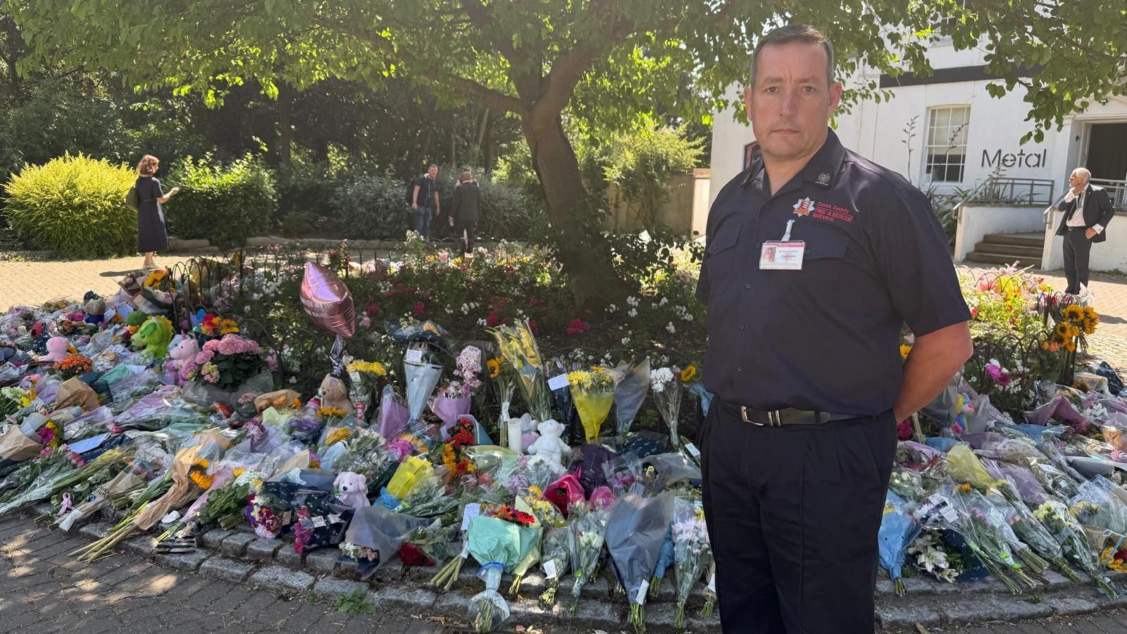 A man standing next to flowers in a park. He has a sombre look on his face 