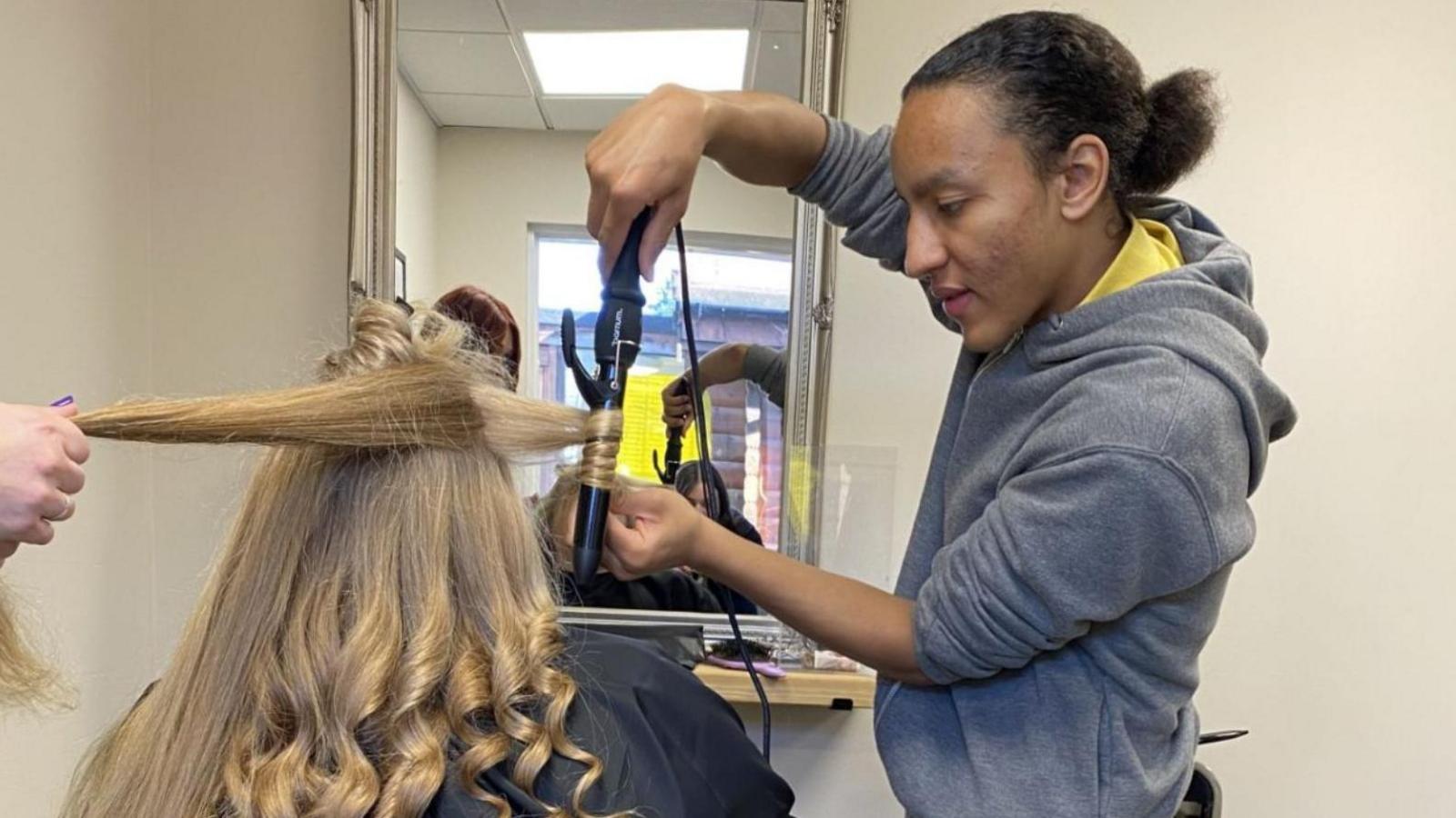 Cameron with dark hair tied at the back wearing a grey top winds hair around some straighteners in front of a mirror. The long blonde hair, with curls at the bottom, belongs to a woman with her back to the camera.