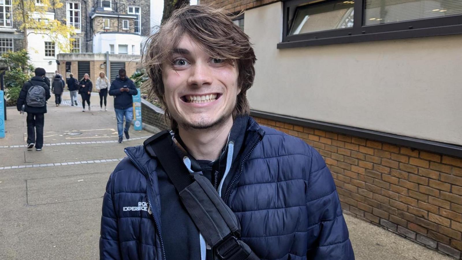 A smiling Gillie Owen with brown hair, wearing a blue puffa jacket and shoulder strap bag