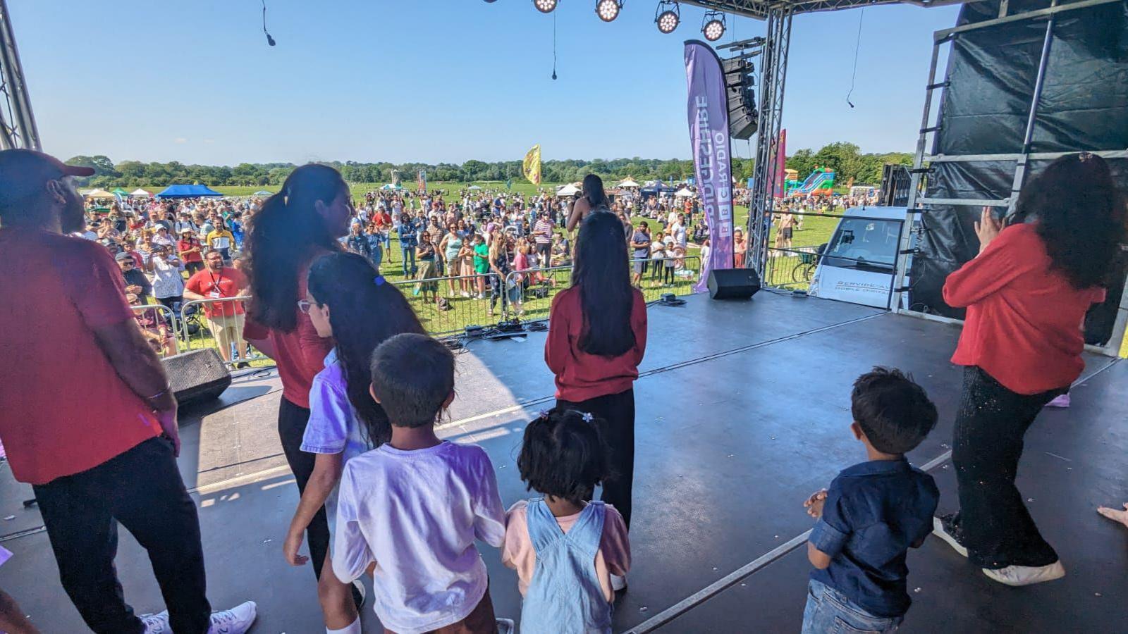 A photograph of children and young people on an outdoors stage, taken from the rear of it - showing the backs of performers and a watching crowd. It is a hot, sunny day, with many people stood on the grass.