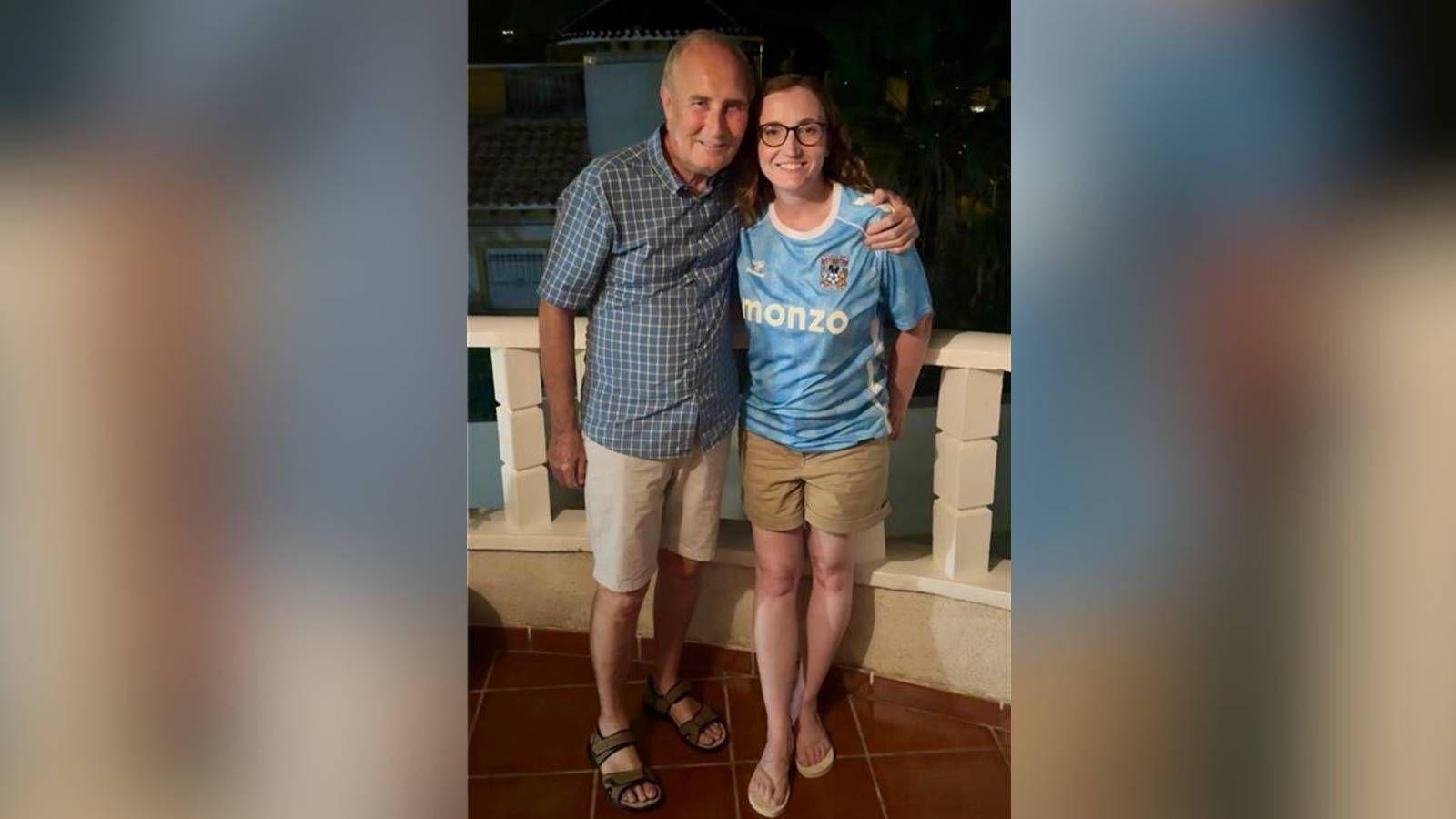 Emma Earnshaw pictured smiling with her dad. She is wearing a Coventry City shirt and khaki shorts while stood next to her dad, who is also smiling, wearing a light blue checked shirt and cream shorts.