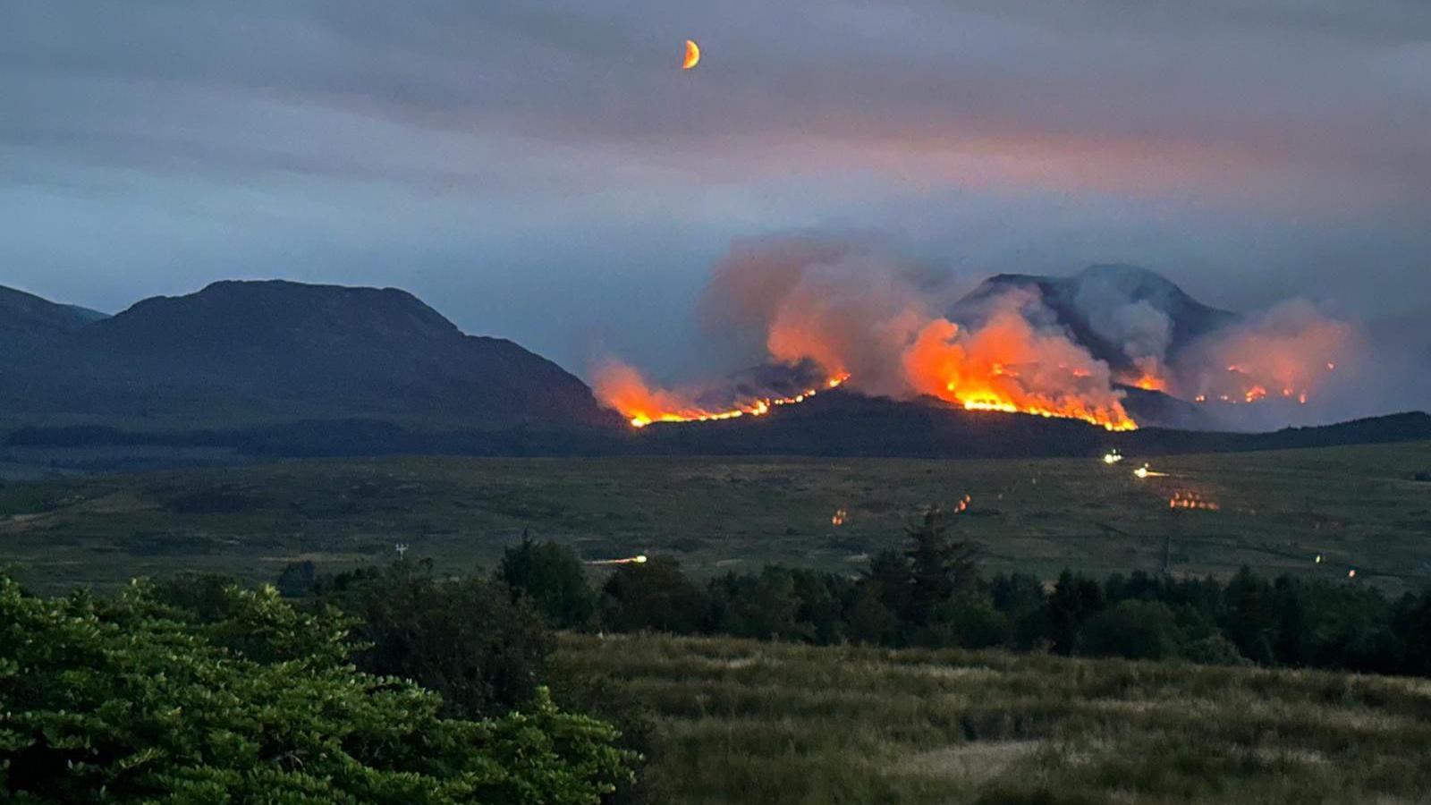 A line of fire on a mountain set against a darkening sky. Fields can be seen in the foreground, with the moon visible in the night sky.