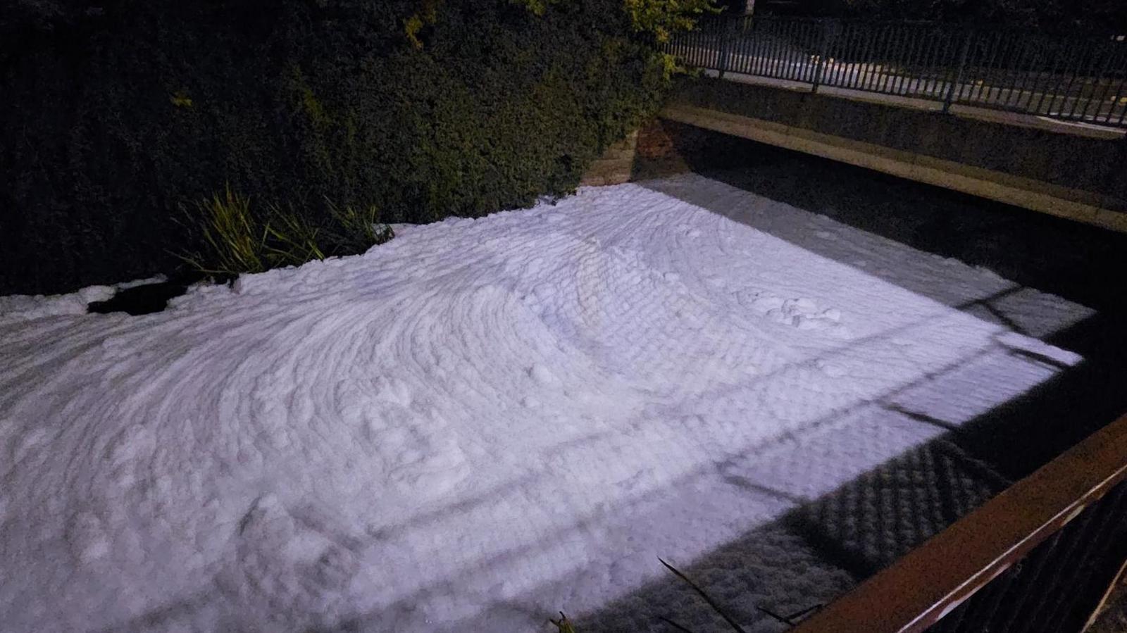 A thick layer of white foam with ripples in it can be seen completely covering the surface of a river which is in the foreground with a bridge in the background.