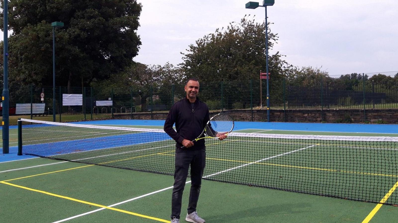Mohammed Rafique is pictured on a refurbished tennis court in Meanwood Park in Leeds. He is wearing trainers and dark athletic clothing and is holding a tennis racquet in his hand