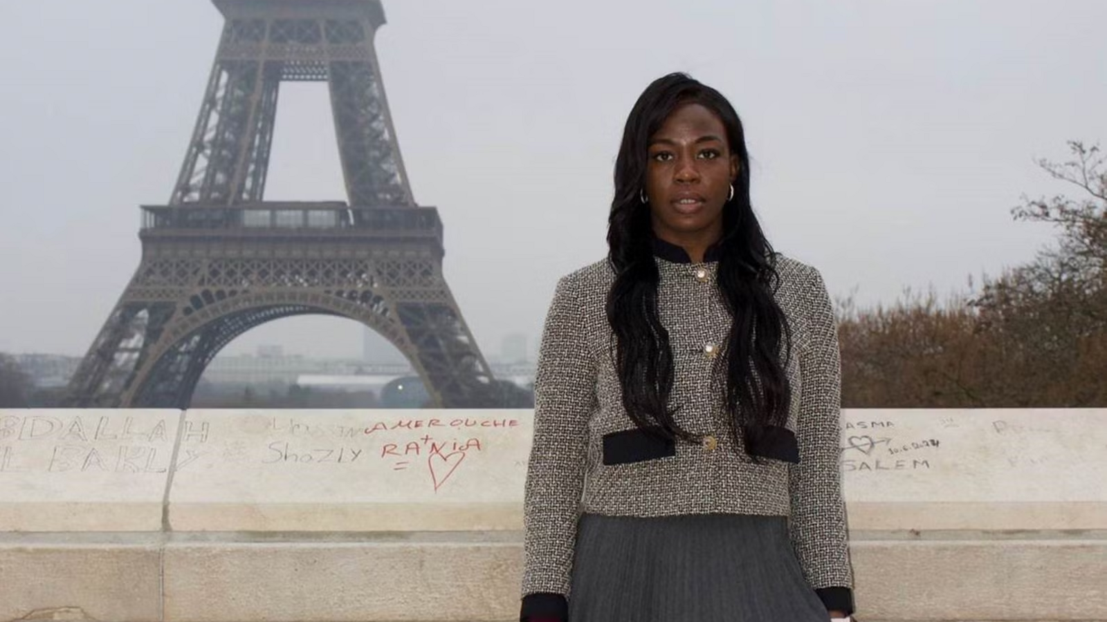 Jamelatu Tsiwah stands by the Eiffel Tower. She is wearing a grey jacket and has long dark hair. 