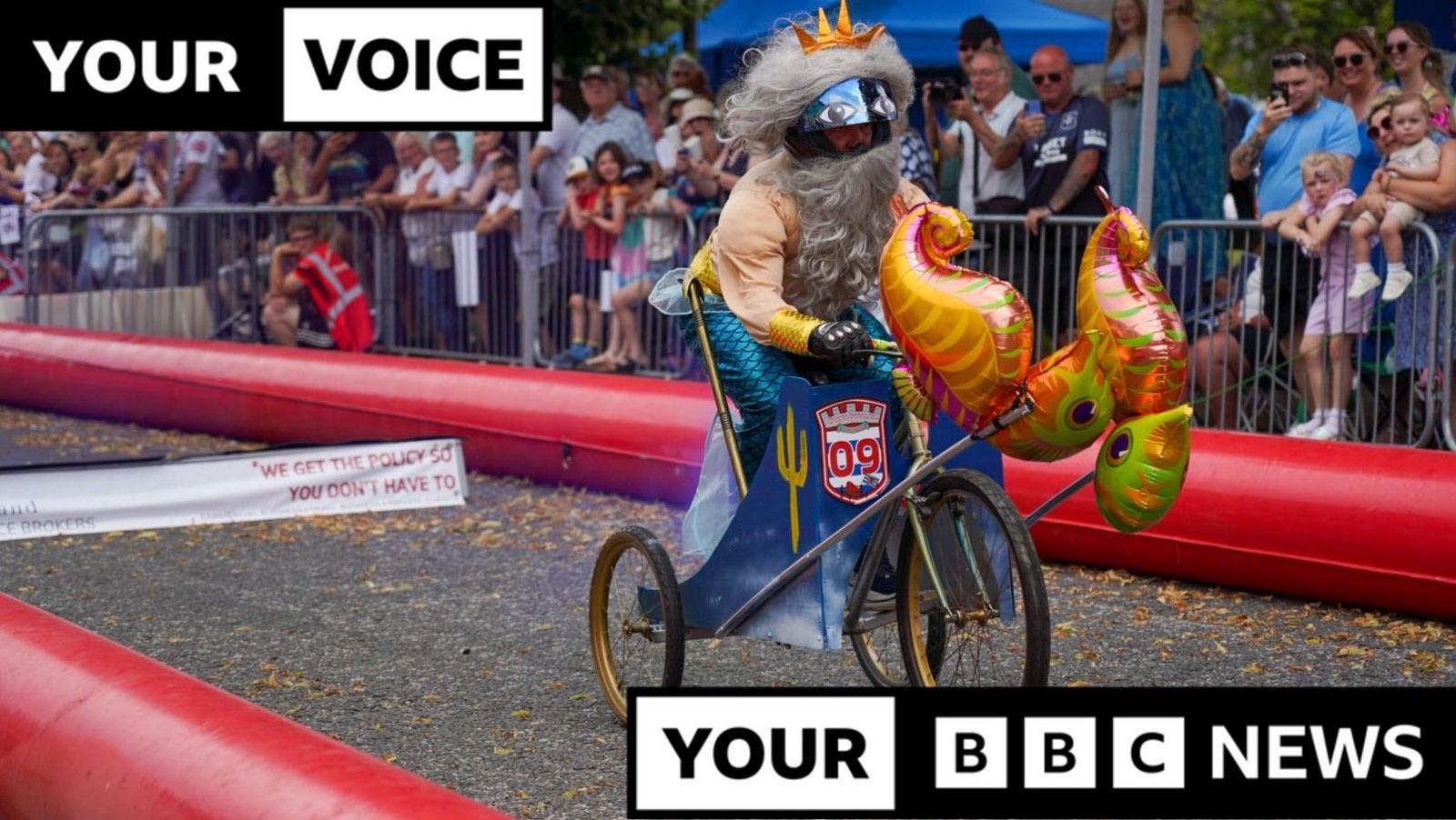 A competitor in Castle Douglas Soapbox Derby. A man dressed as a mermaid is riding a three-wheeled bike along the course. A crowd of spectators is watching from behind a barrier.