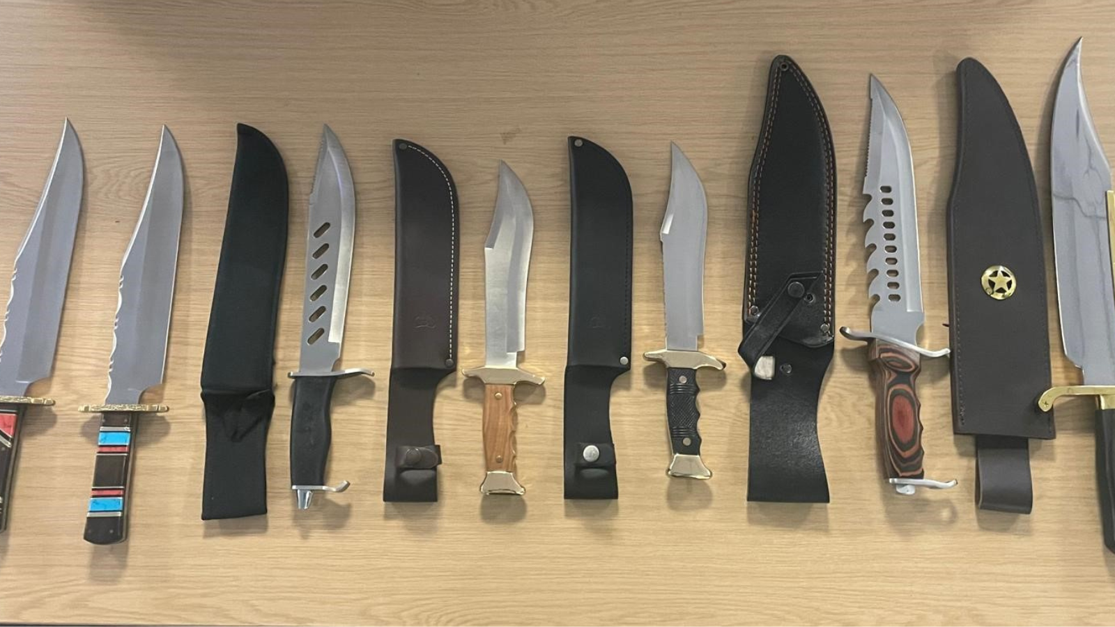 Twelve knives are laid out on a wooden desk.