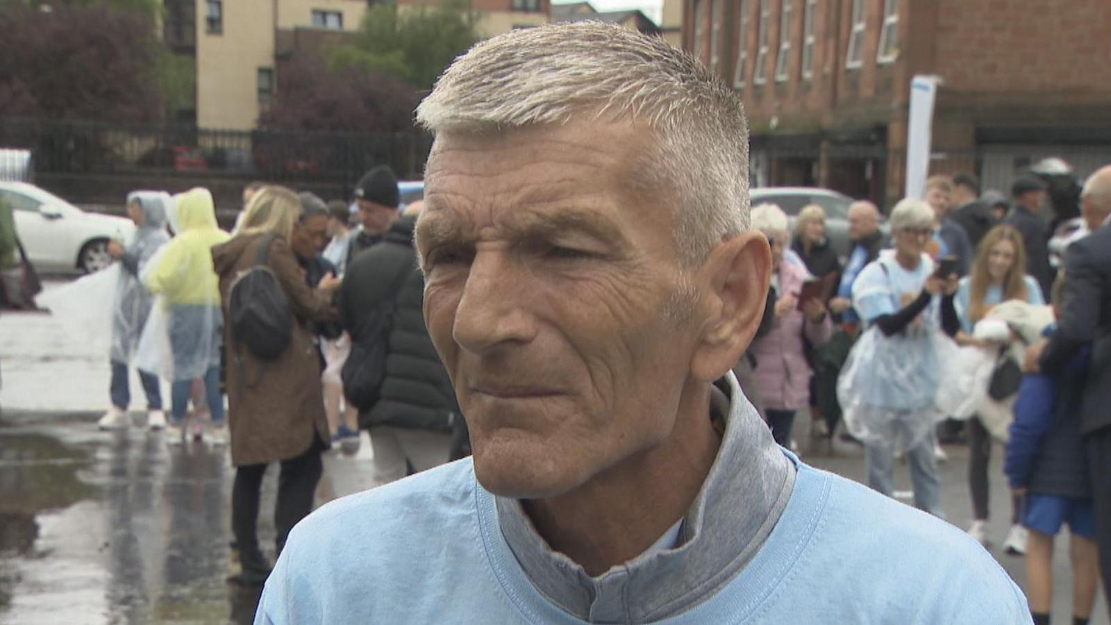 Neil McCrimmon looking slightly off camera. He has grey hair and is wearing a light blue t-shirt over a grey jumper.