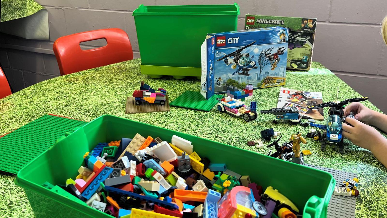 A table with Lego plastic building bricks. A child is building a helicopter. Lego-built cars are on the table.
