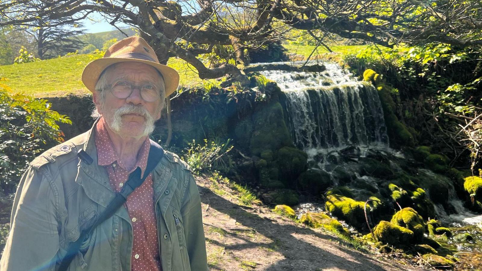 Doug Chalk posing next to the rocky waterfall at Littlebredy. It's a bright sunny day and he is wearing a mustard colour trilby hat, a plum collar shirt with small white spots and a khaki green jacket. He has a large grey moustache and round glasses.
