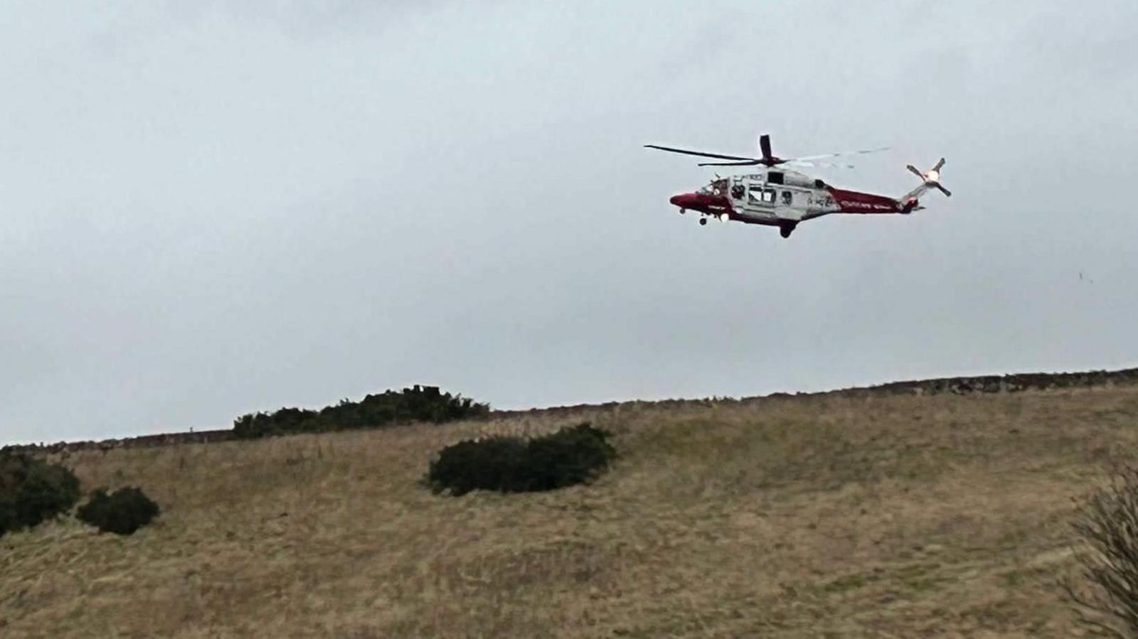 A red and white helicopter hovers in a grey sky above a field. Four bushes below and yellow-brown grass.