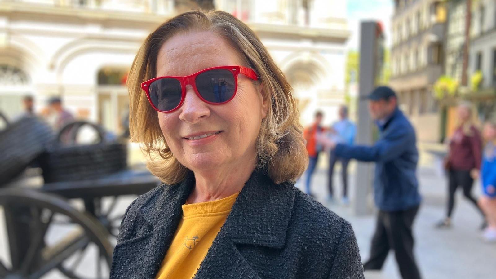 Clodagh Keating has red glasses, a yellow top and a navy coat. She's got blond hair, worn short and smiles at the camera