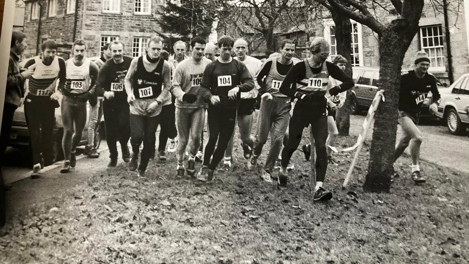 A black and white photograph of lots of men wearing bibs and just beginning to run 