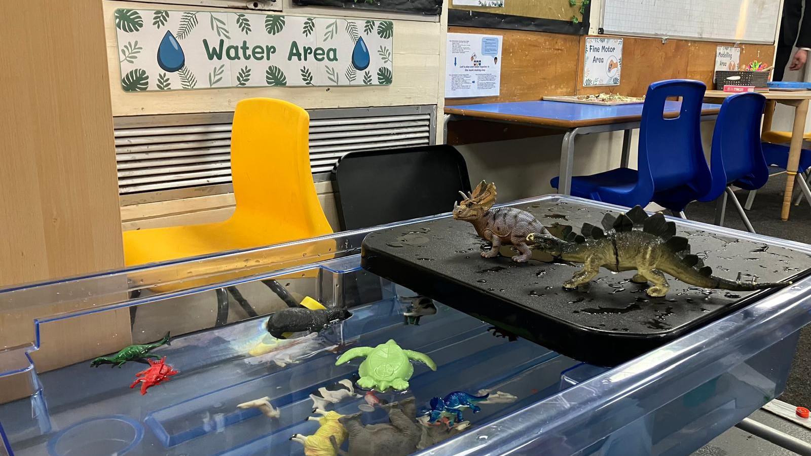 A tank of water with dinosaurs floating inside. A sign behind the tank says "water area" chairs and tables are in the background.