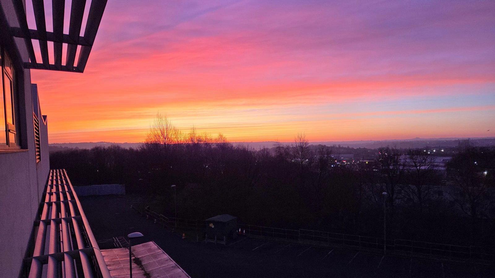 A view from a flat window of the rays of sunrise over a town with trees dotted throughout. The sky is red, yellow, blue and orange.