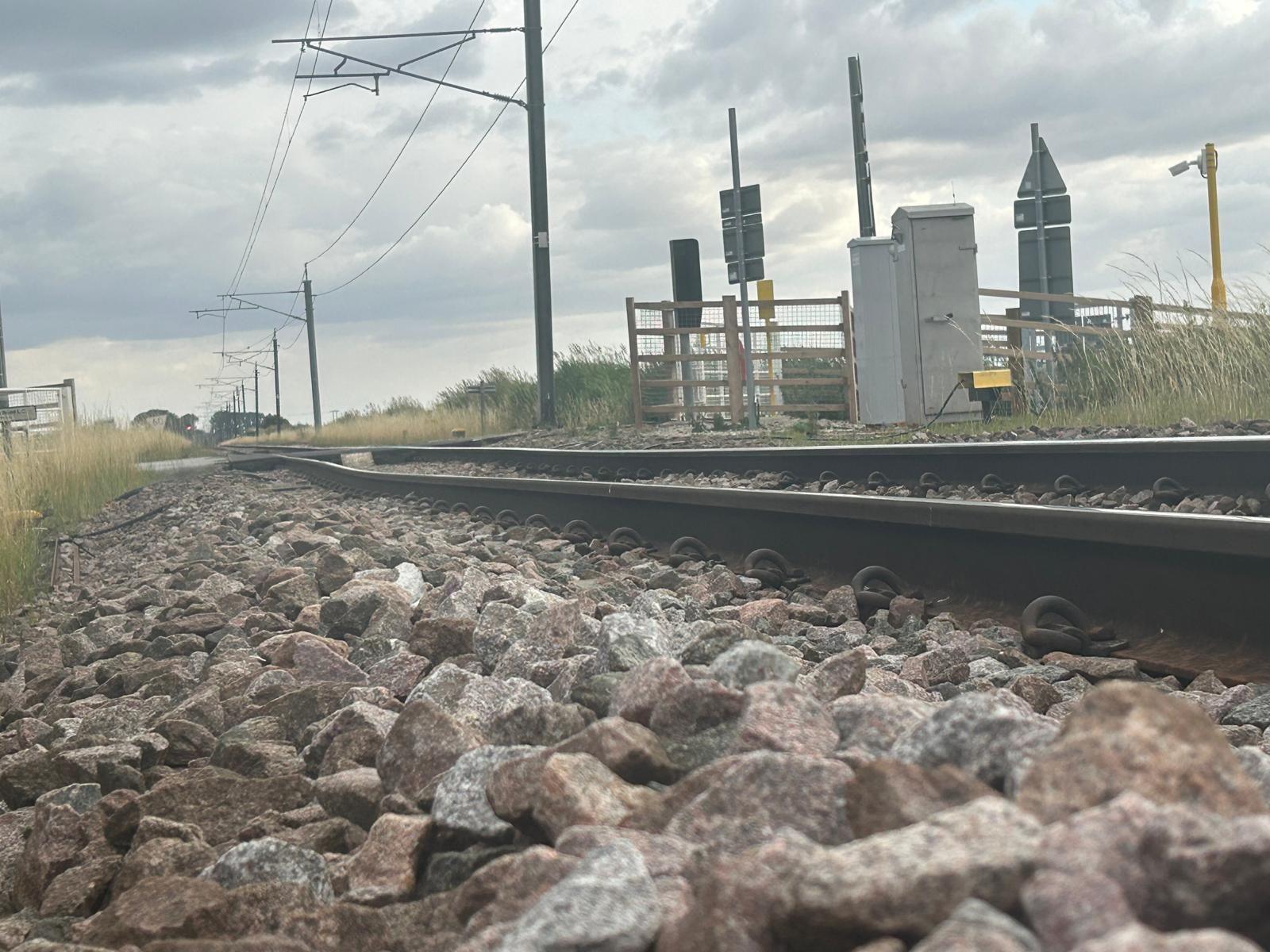 An uneven piece  of railway track with ballast or large stones alongside it. Overhead lines and a level crossing can be seen too with signage and fencing. Dry tall grass grows alongside the track. 