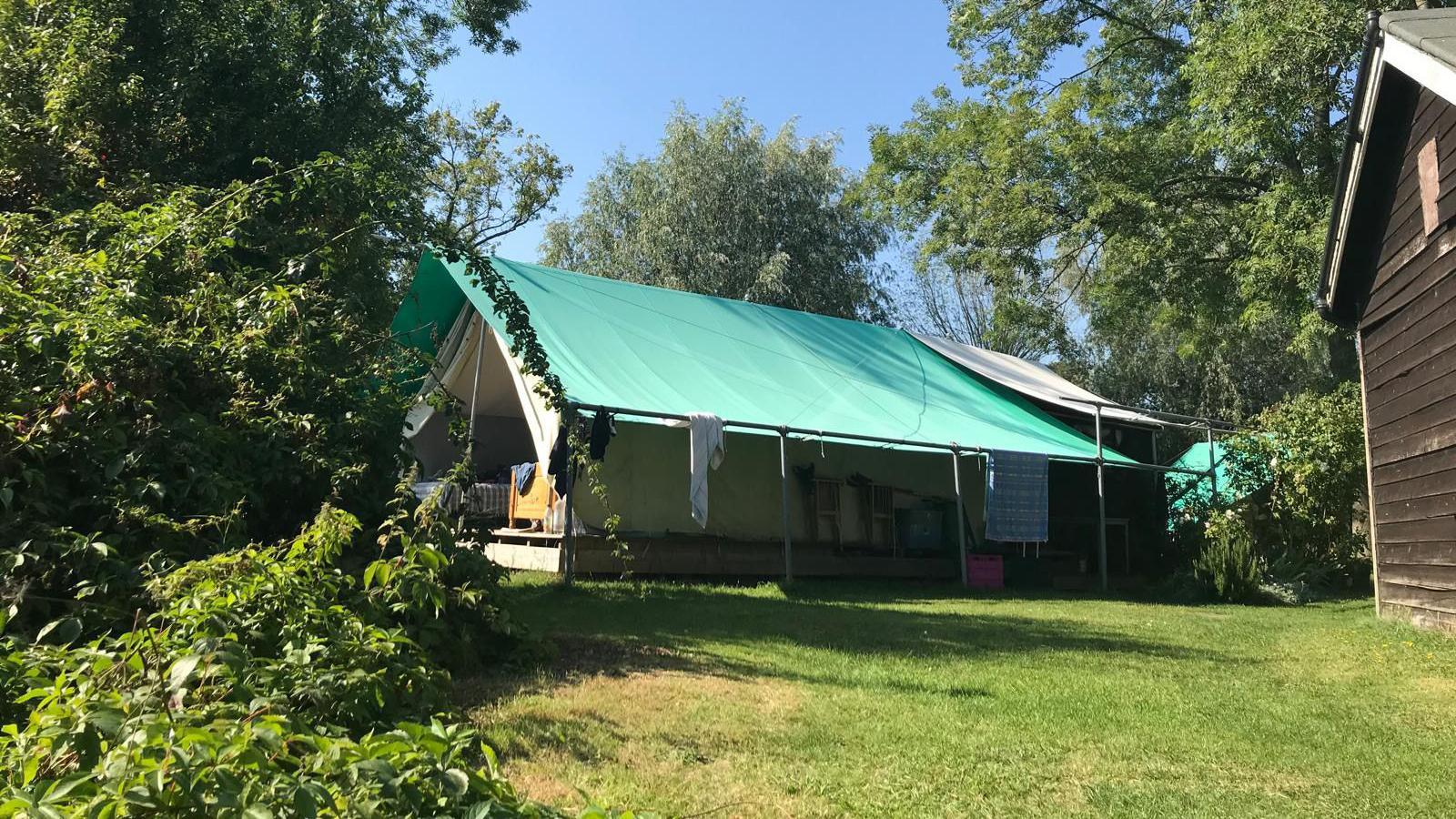 A large canvas tent structure is pictured in a pleasant field, with a green roof and clothes hanging from the side. A hut can be seen on one side and a bush on the other. 