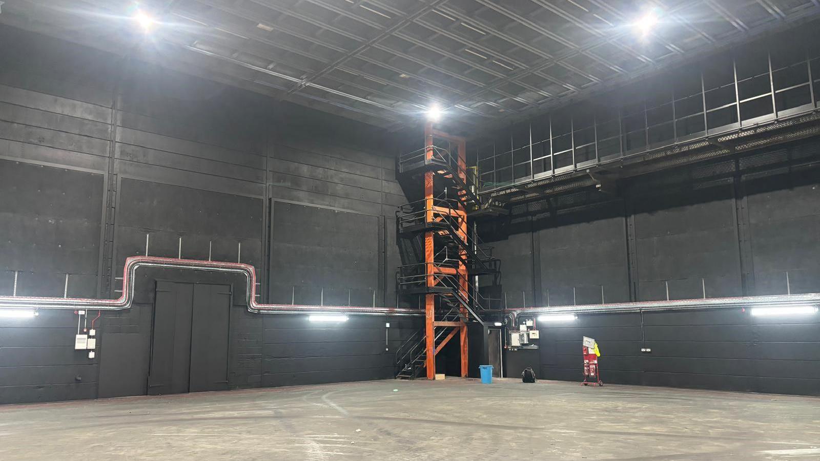 The inside of Stage 7, a large warehouse like room with lighting on the ceiling and around the sides. In one corner is a three storey open staircase with orange pillars.