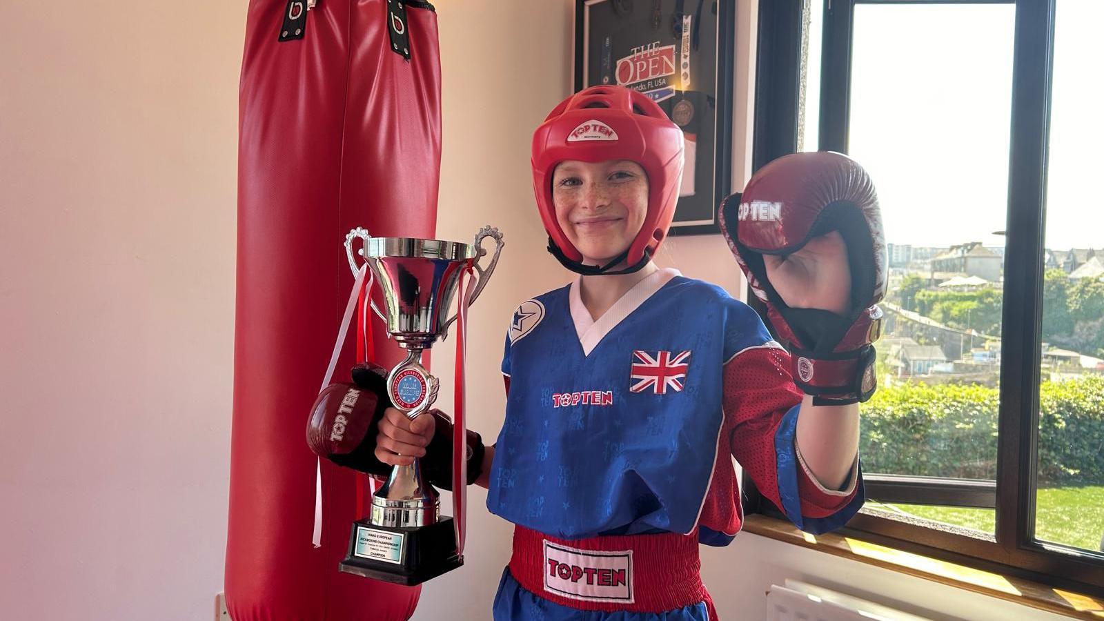 Anya is stood in her kickboxing gear by a punch bag holding a trophy