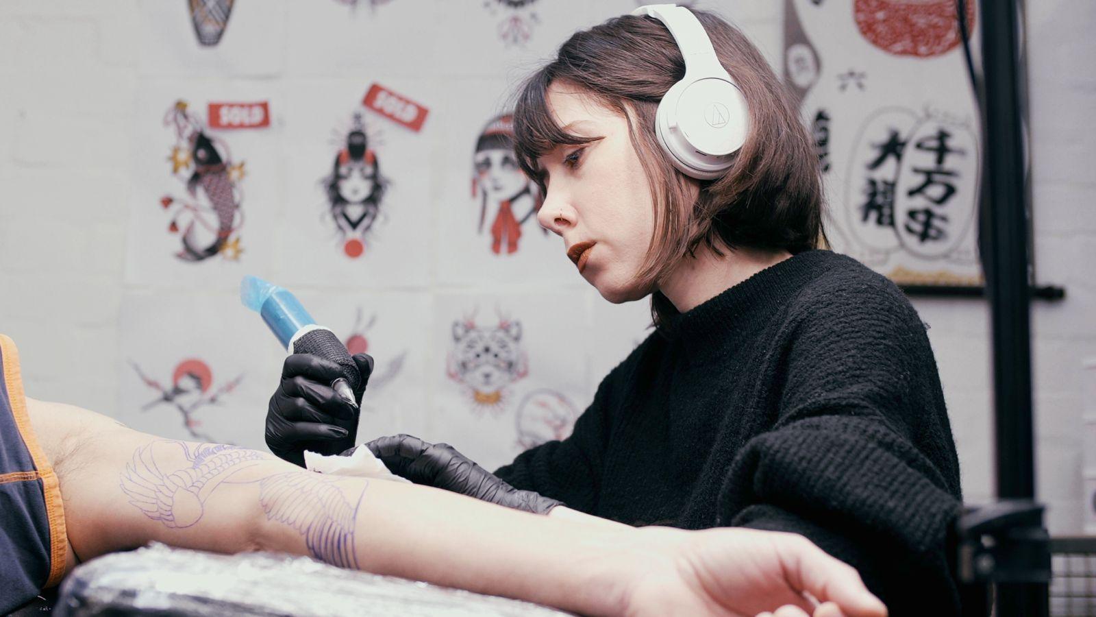 Loiz leaving over a client's arm and tattooing. She has brown hair tied half up, and is wearing black latex gloves, and is holding a tattoo gun