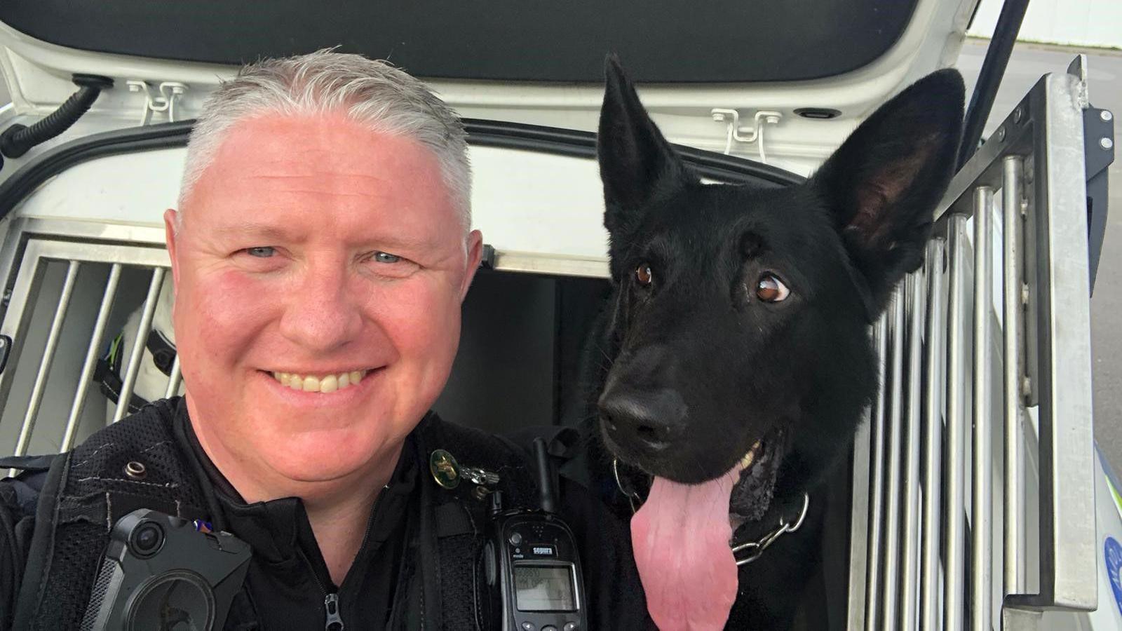 A police officer smiling at the camera with a black German shepherd, who is sticking his tongue out