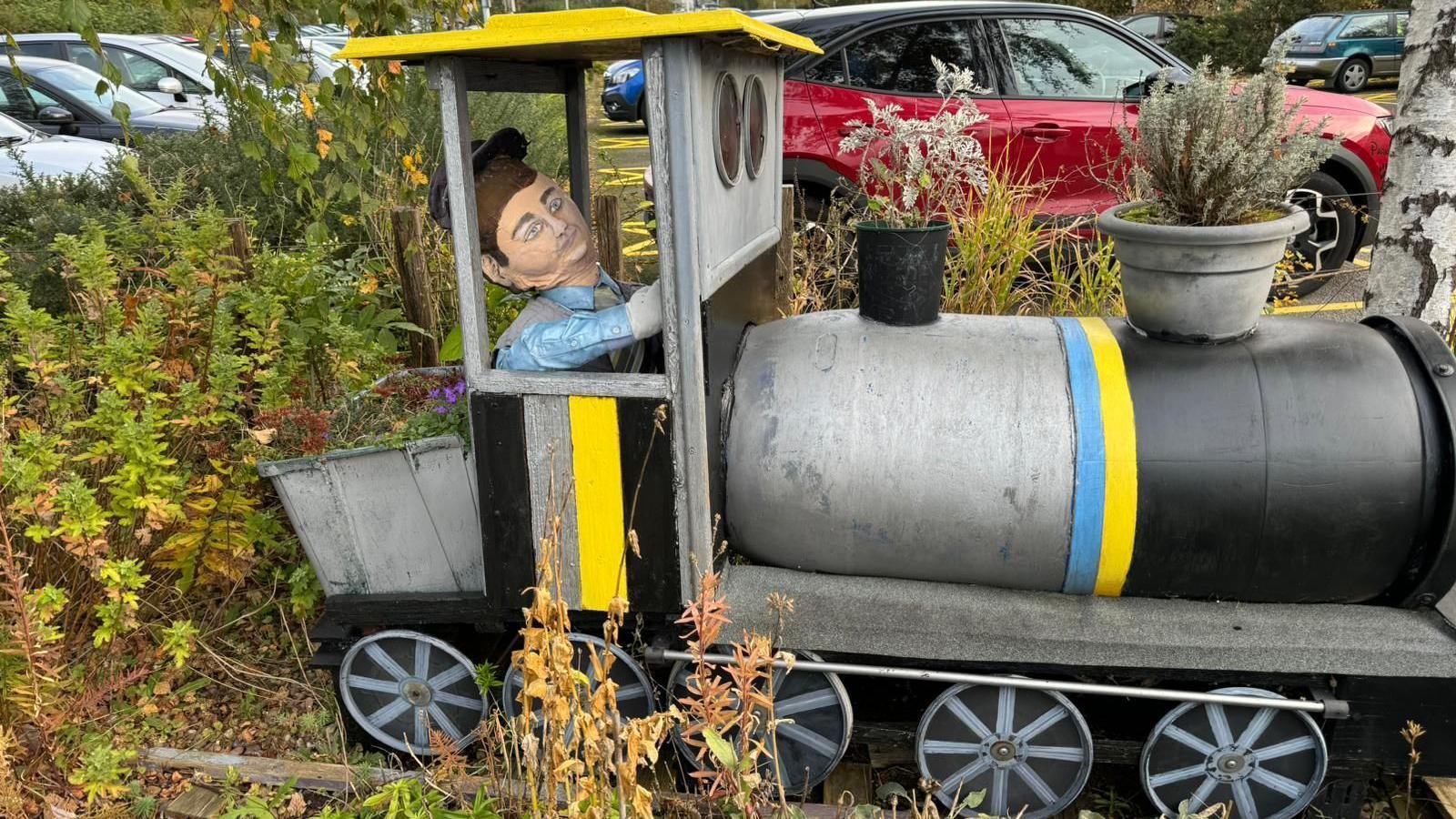 A grey, black and yellow model of a train with a stuffed figure in the cabin and plantpots for a funnel