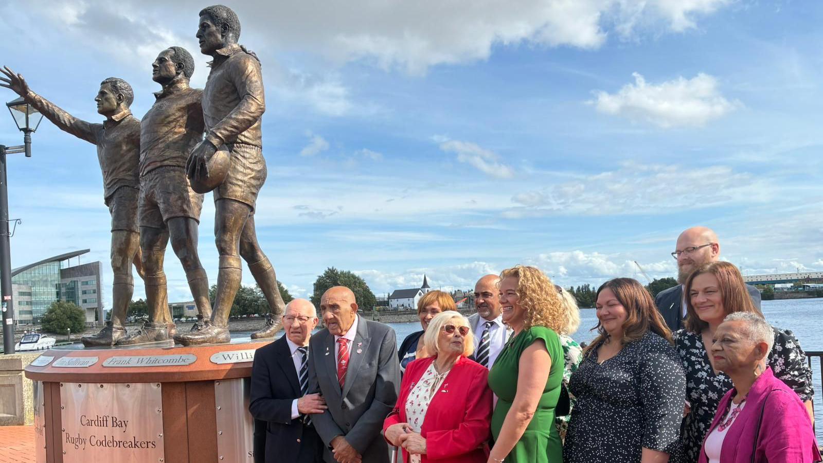 On the left is Welsh rugby "codebreakers" statue. On the right, Billy Boston stands with his family and members of the Codebreakers Committee as photos are taken at the unveiling in Cardiff Bay. The man closest to the statue has a dark blue jacket with a white shirt and striped tie, next to him is Sir Billy with a dark grey suit and red tie. There is a woman peaking out behind him with ginger hair, and next to him is a balding man with a white shirt and black and white striped tie. In front of him is a woman with a red jacket and white shirt with a bright blonde bob and shades, she looks at a woman with curly blonde hair who's wearing a green dress and smiling. Next to her is a woman with shoulder-length dark hair and a dark blue dress, next to her is a woman with short grey hair and a pink jumper and light pink top. Just behind her is a woman with a dark blue dress and dark brown hair and behind her is a balding man with a beard. The bay of Cardiff can be seen behind them.