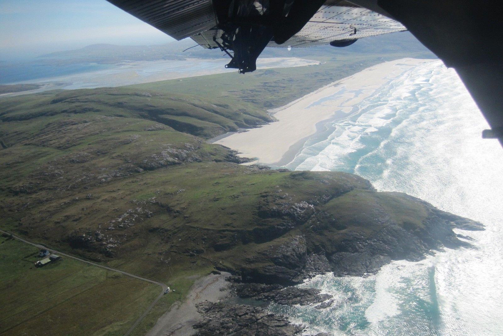 Picture taken from a plane, showing a small piece of the wing with an island below, surrounded by a sandy beach with white waves splashing on the shore