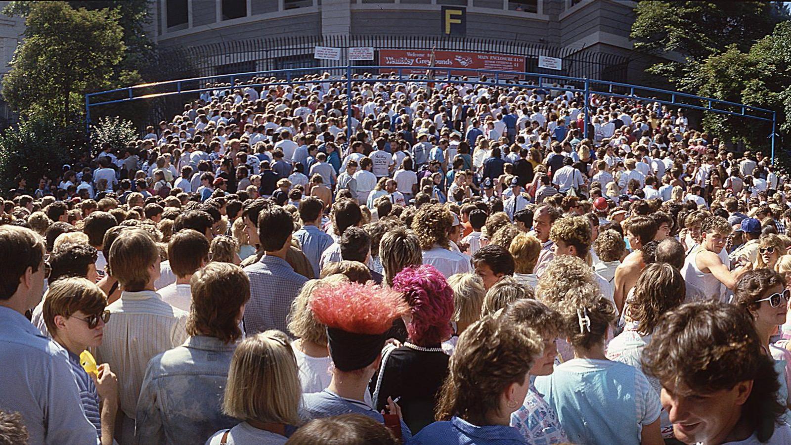 Crowds waiting outside the stadium entrance under bright daylight and filing up steps toward a gate beneath a “Wembley Grounds” sign.