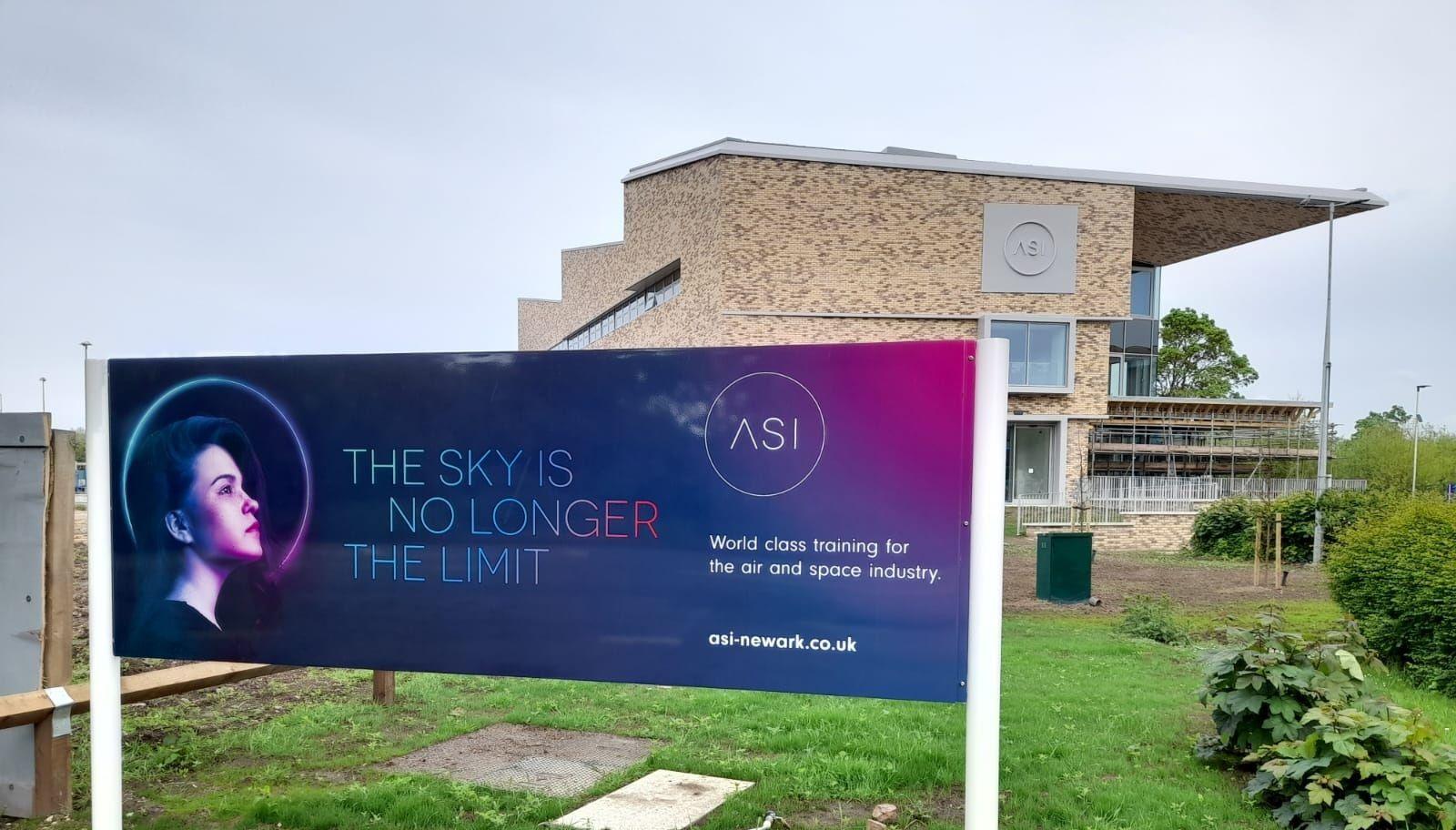 A sign for the ASI stands in front of the light-brown brick building. It has a roof that protrudes beyond the building below it. Some scaffolding is visible in the background.