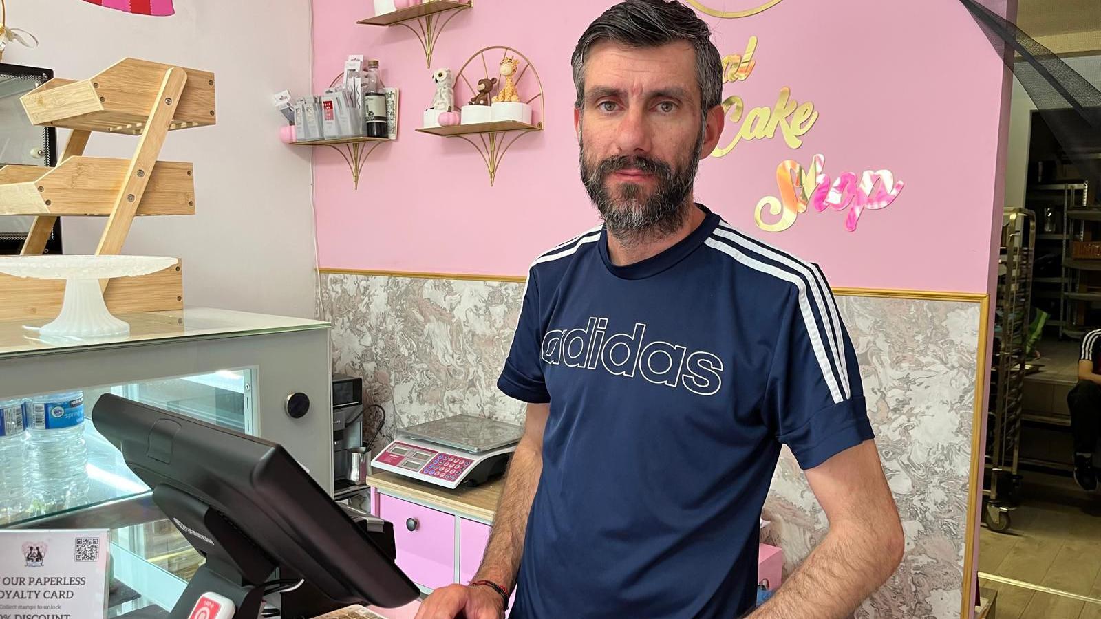 Constantin Manzu, who is 40 and has dark hair and a beard with streaks of grey, stands behind the counter at his cake shop wearing a blue Adidas branded t-shirt.