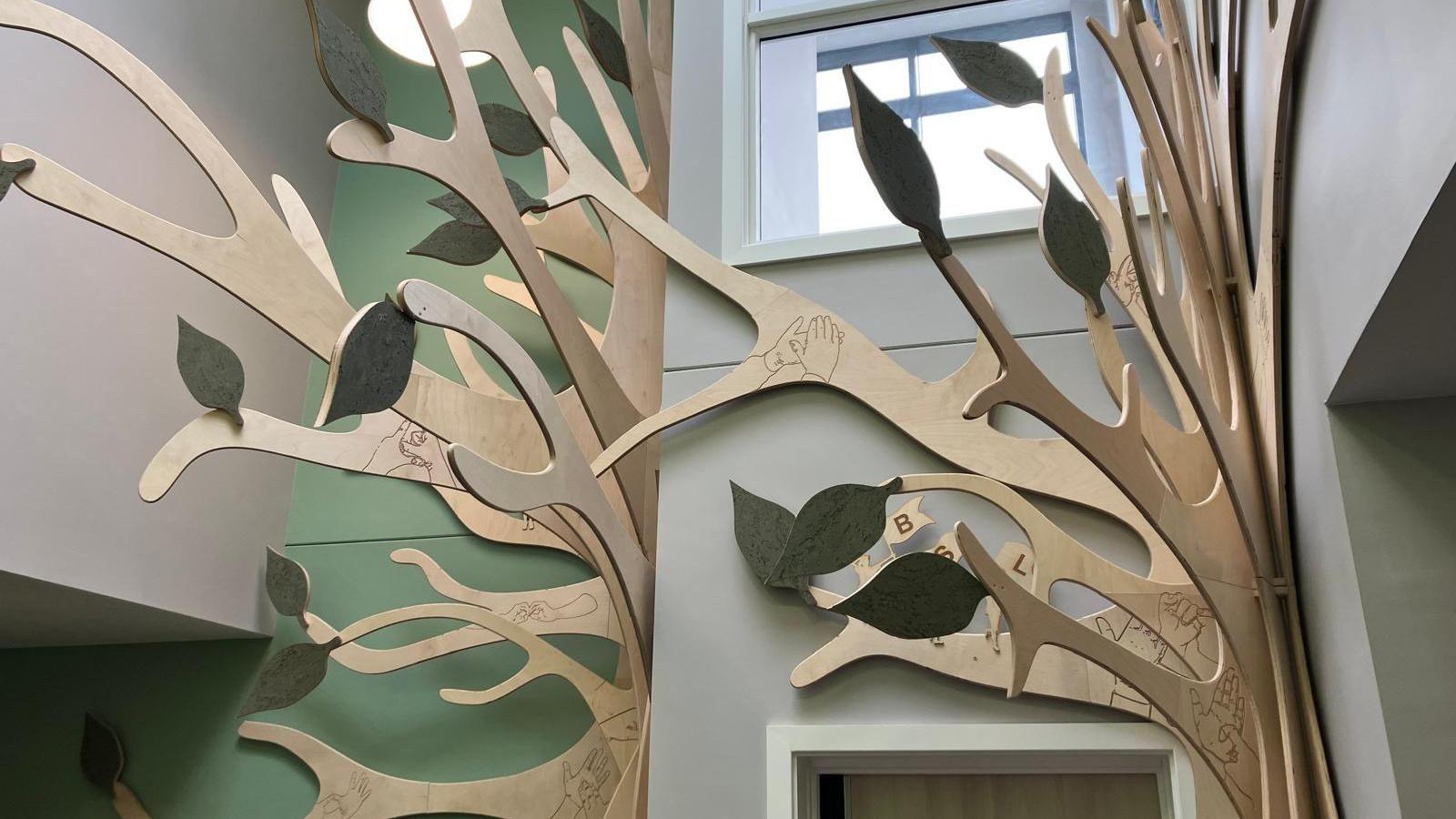 The top part of the tree sculpture rising up to the ceiling of the school's reception. You can see an upper window filled with light behind its branches. The walls behind it are grey and green.
