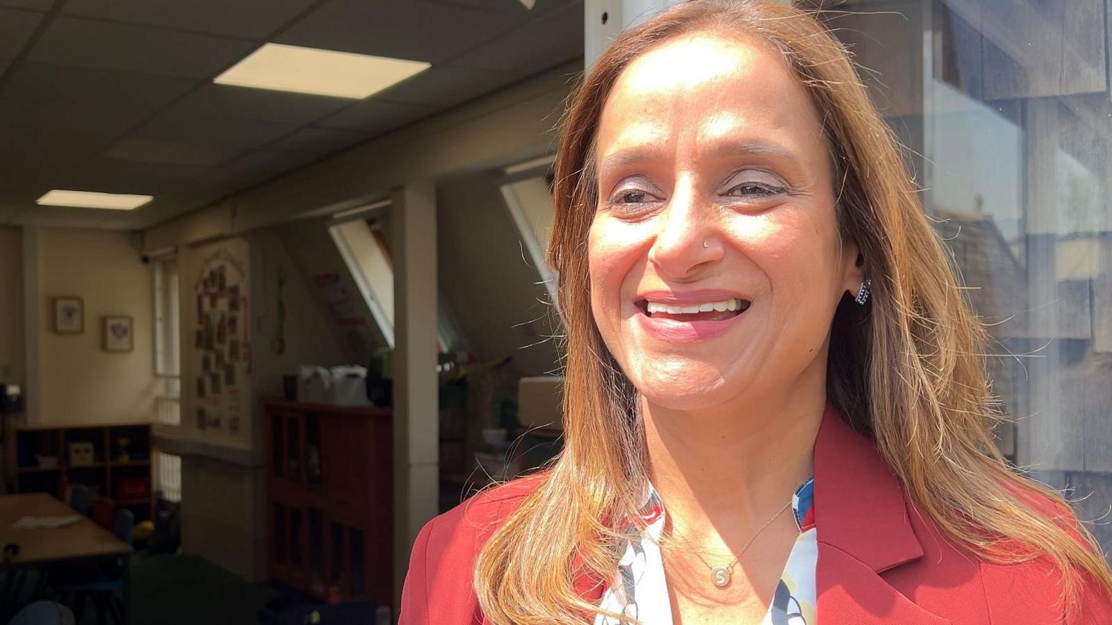 Satinder Bell has long brown hair which is around her shoulders. She is smiling, she is wearing a pink blazer and white shirt with blue, red and yellow patterning.