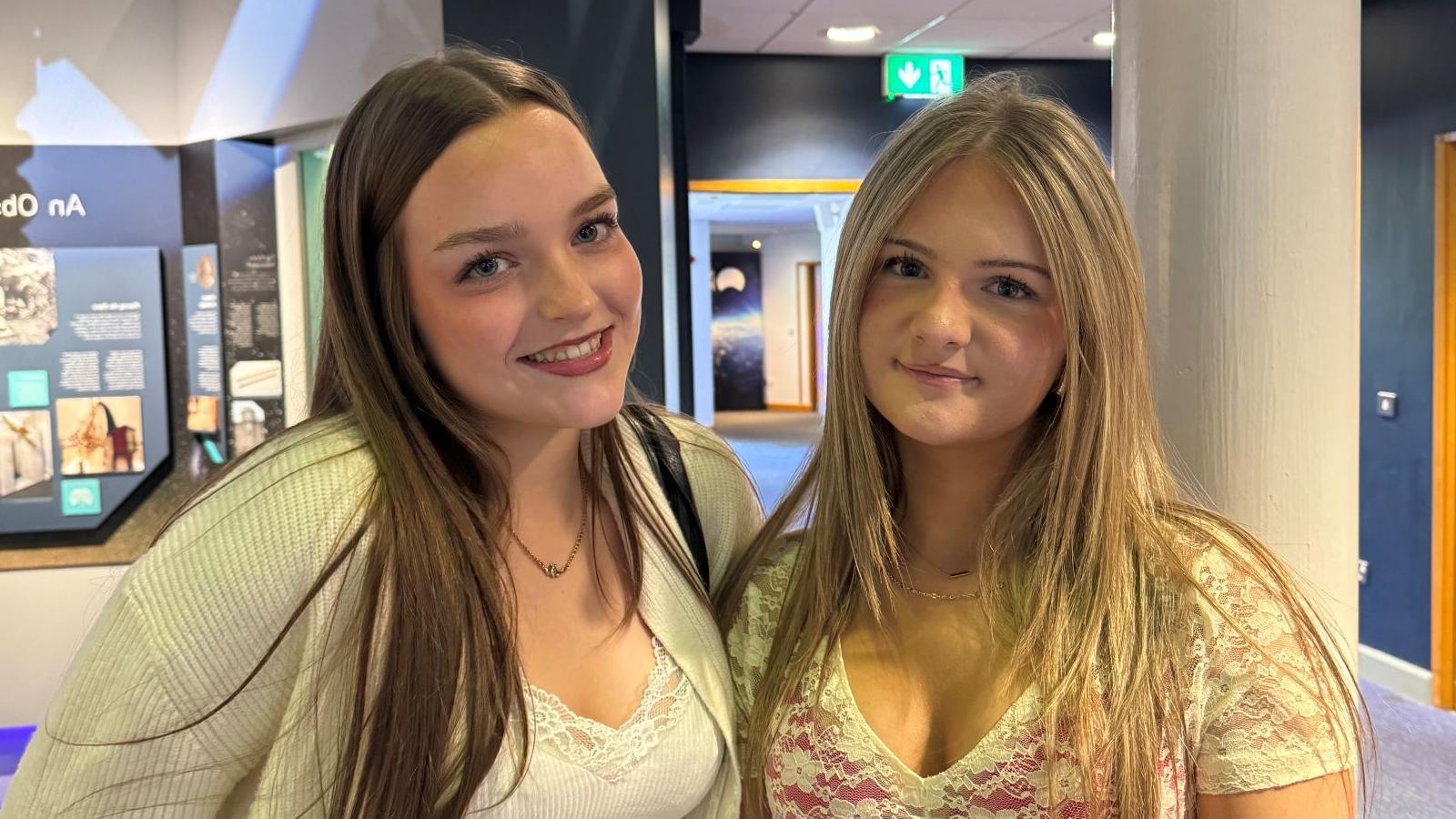 Emma and Tara are two young girls who are standing side by side. Emma on the left has long, dark hair and Tara on the right has long, blonde hair. Both girls are smiling at the camera.