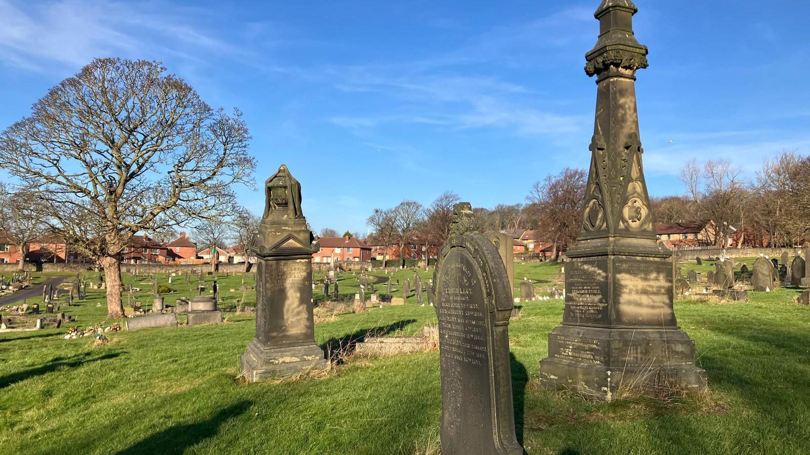 Dewsbury Cemetery. Three ornate gravestones centre in the image. Thousands of graves are in the background as well as a large tree.