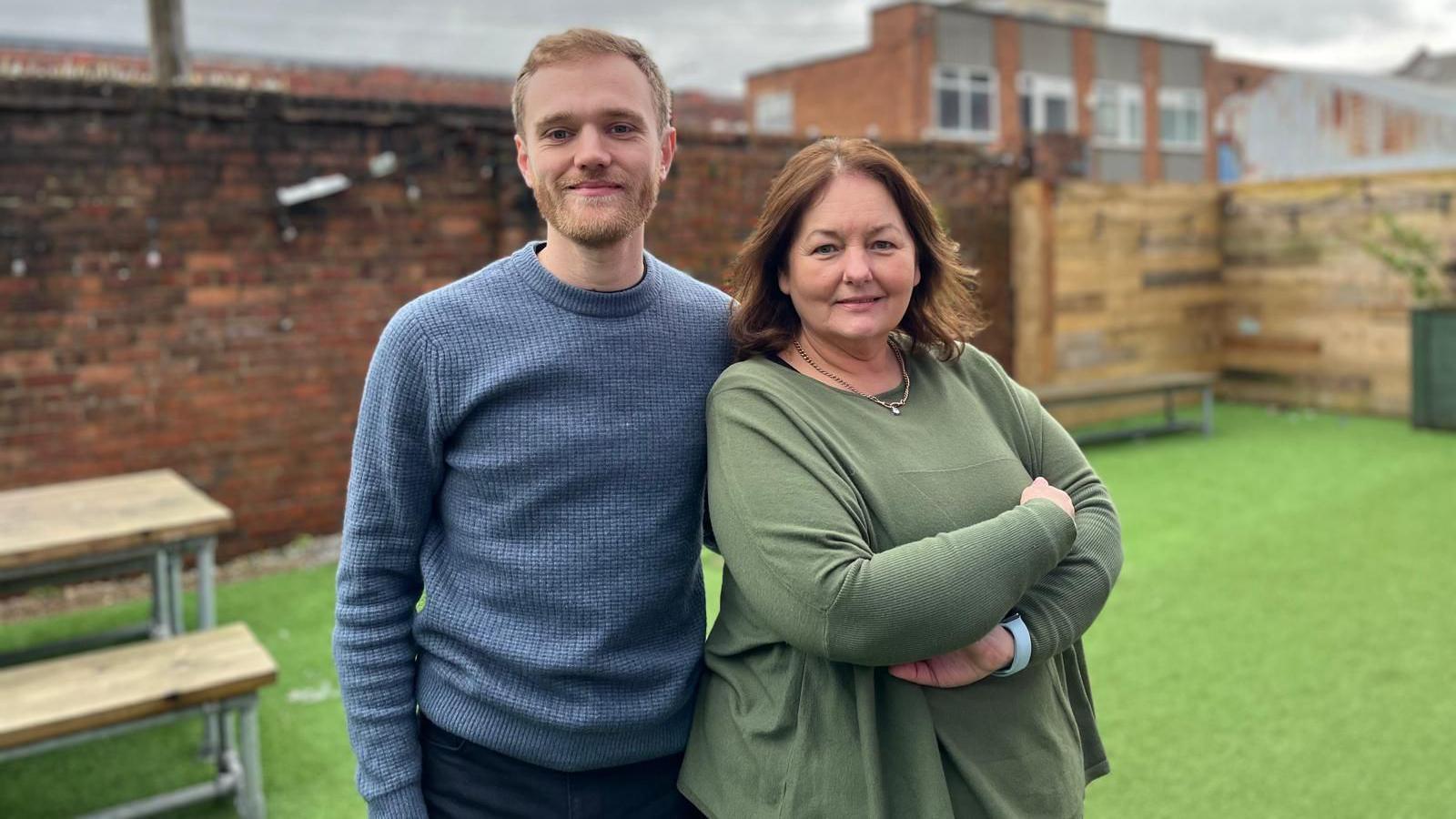 Joseph Burns with sandy coloured hair and a light beard and wearing a blue sweatshirt, and Julie Maddocks with brown hair and a green top, smile at the camera in a beer garden area with tables and benches in the background