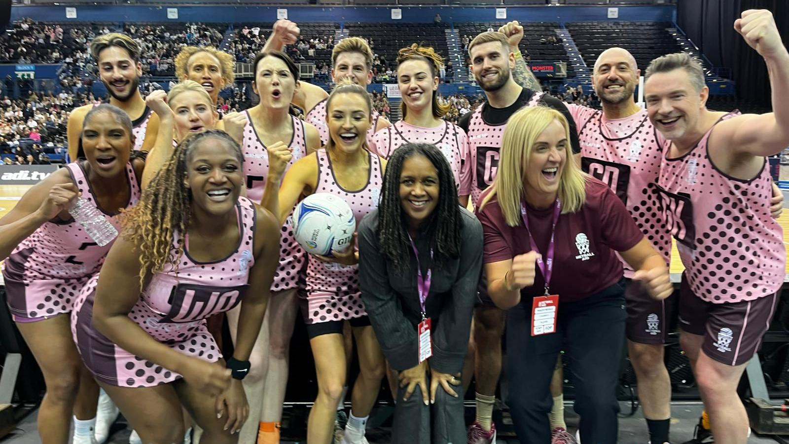 About 14 people dressed in pink netball kit. They are all posing for the camera. They are mix of ages and abilities. We can see they are in an arena. There are hundreds of people in stands behind them.