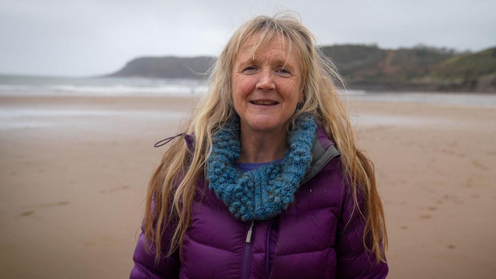 Katie Aviss has long blonde hair and is stood on a beach wearing a purple coat and a blue woolly snood