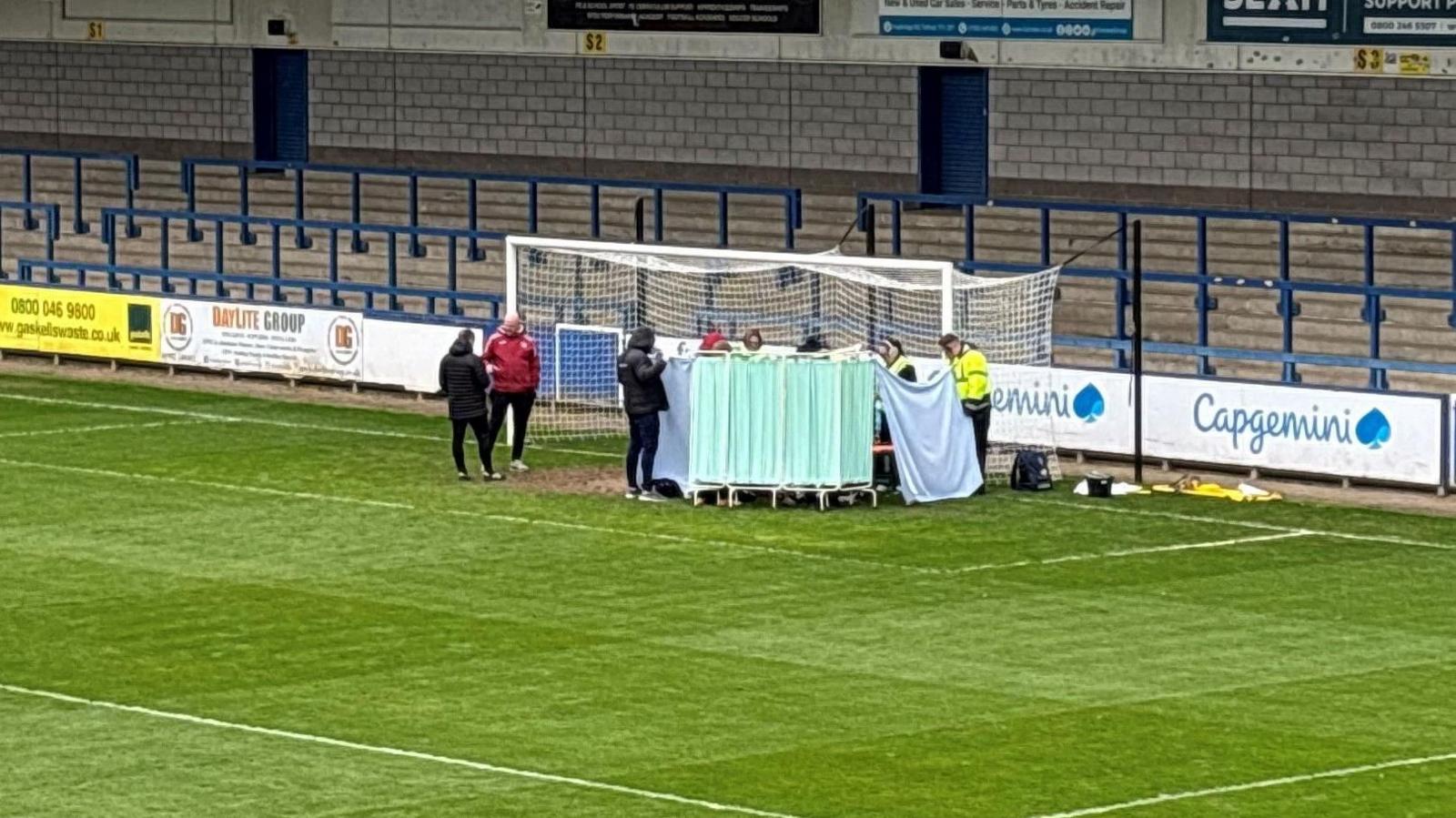 A green medical screen stands in front of the goal at a football stadium. Staff hold up blue sheets on either side to protect the player's privacy.  