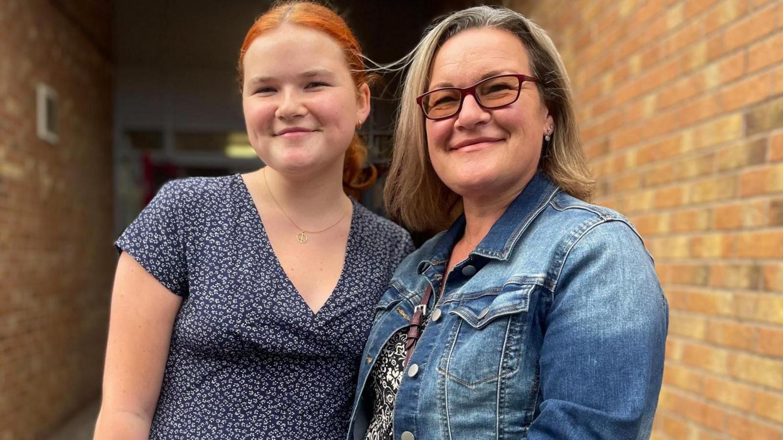A teenage girl with red hair stands next to her Mum who wears a denim jacket