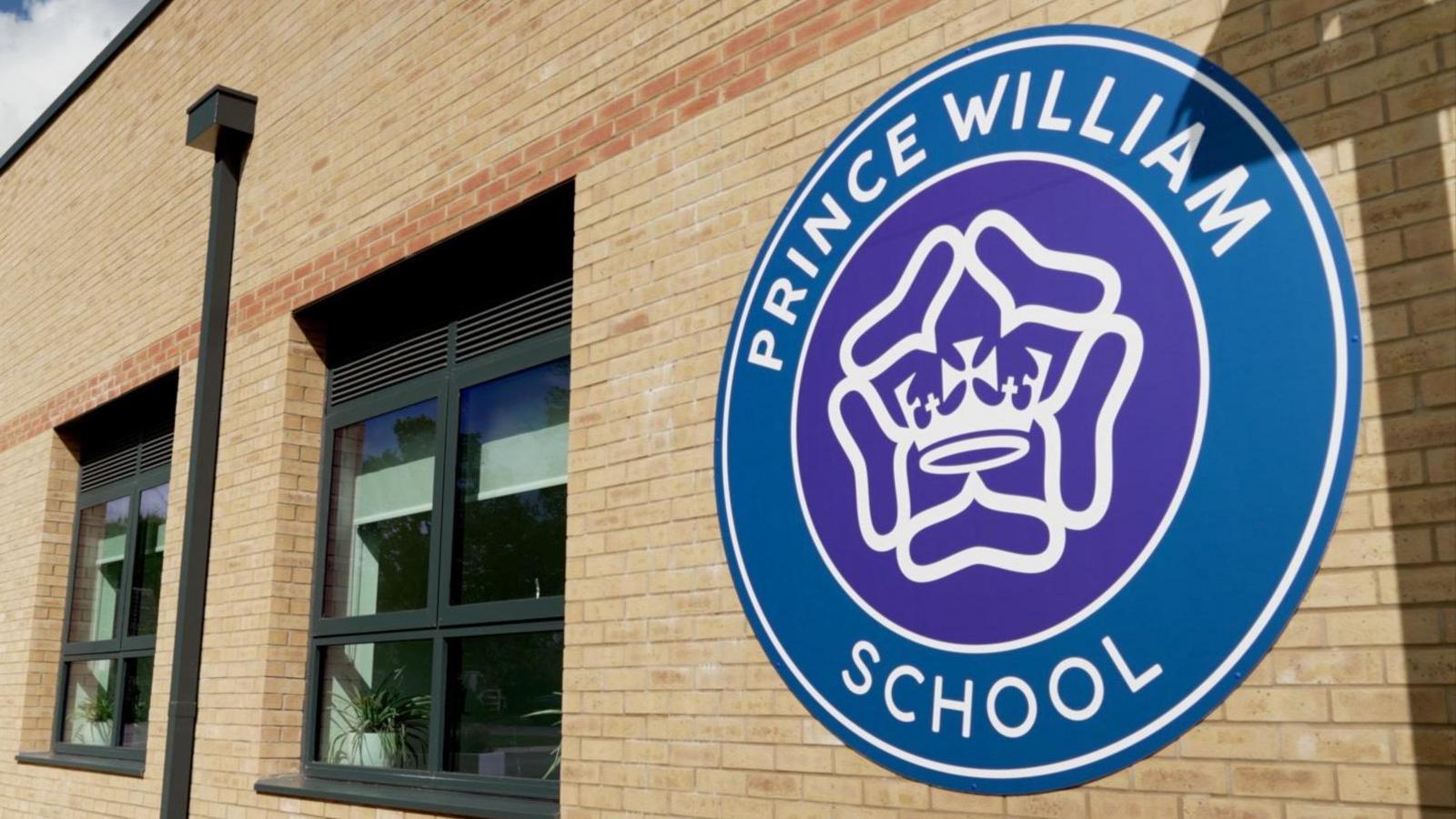 An external brick wall with a large circular Prince William School logo. The middle of the logo is a purple circle, which has a white crown and white rose-style motif in the middle. The words "Prince William School" are written in white against a blue background, which runs around the central circle. Two windows and a drainpipe can be seen further along the wall in the background.