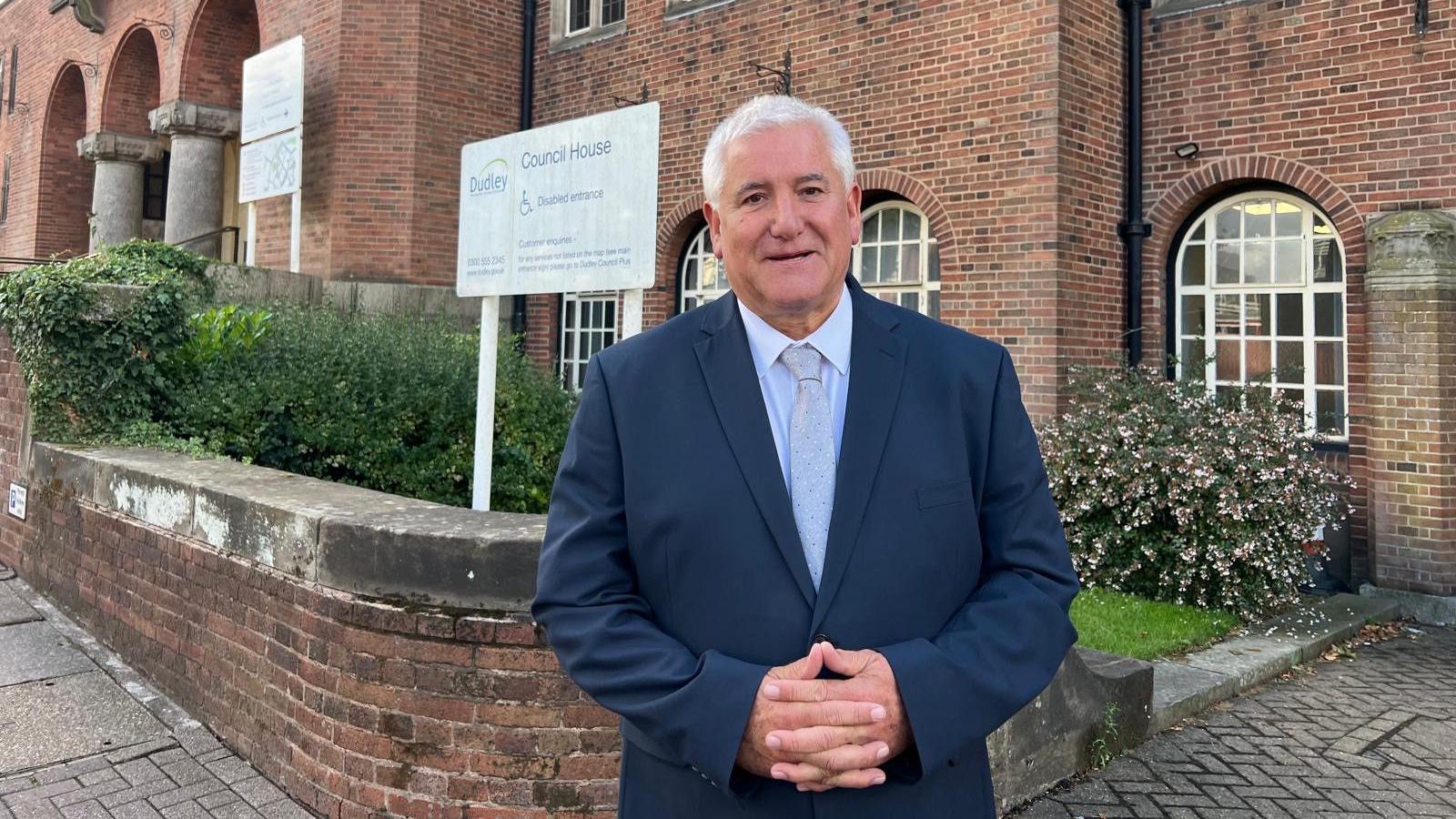 A man wearing a navy suit, grey tie and white shirt and with his arms crossed is standing in front of a brick building, a wall and bushes. There is a sign behind him which reads Dudley Council House.
