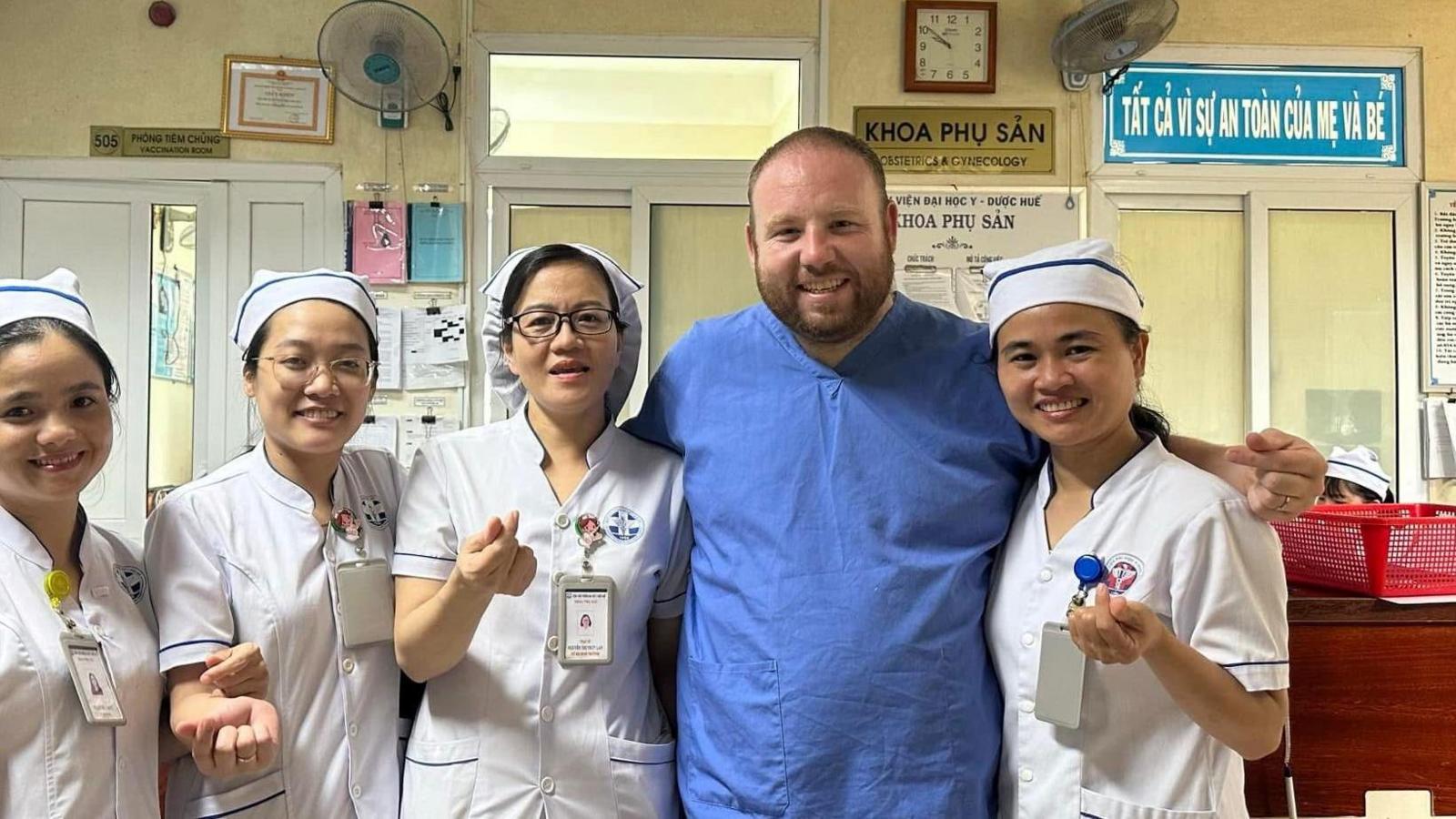 Male Midwife Lee Gigg, smiling, on his elective, before losing weight, wearing blue hospital scrubs, standing alongside four smiling Vietnamese female midwifery staff wearing white uniforms with blue piping and nurses hats.